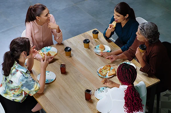 Happy cheerful business woman group team talk gossip eat pizza share lunch together indoor office work place. Indian diverse female sitting around table enjoy break fast tasty sandwich have fun joy