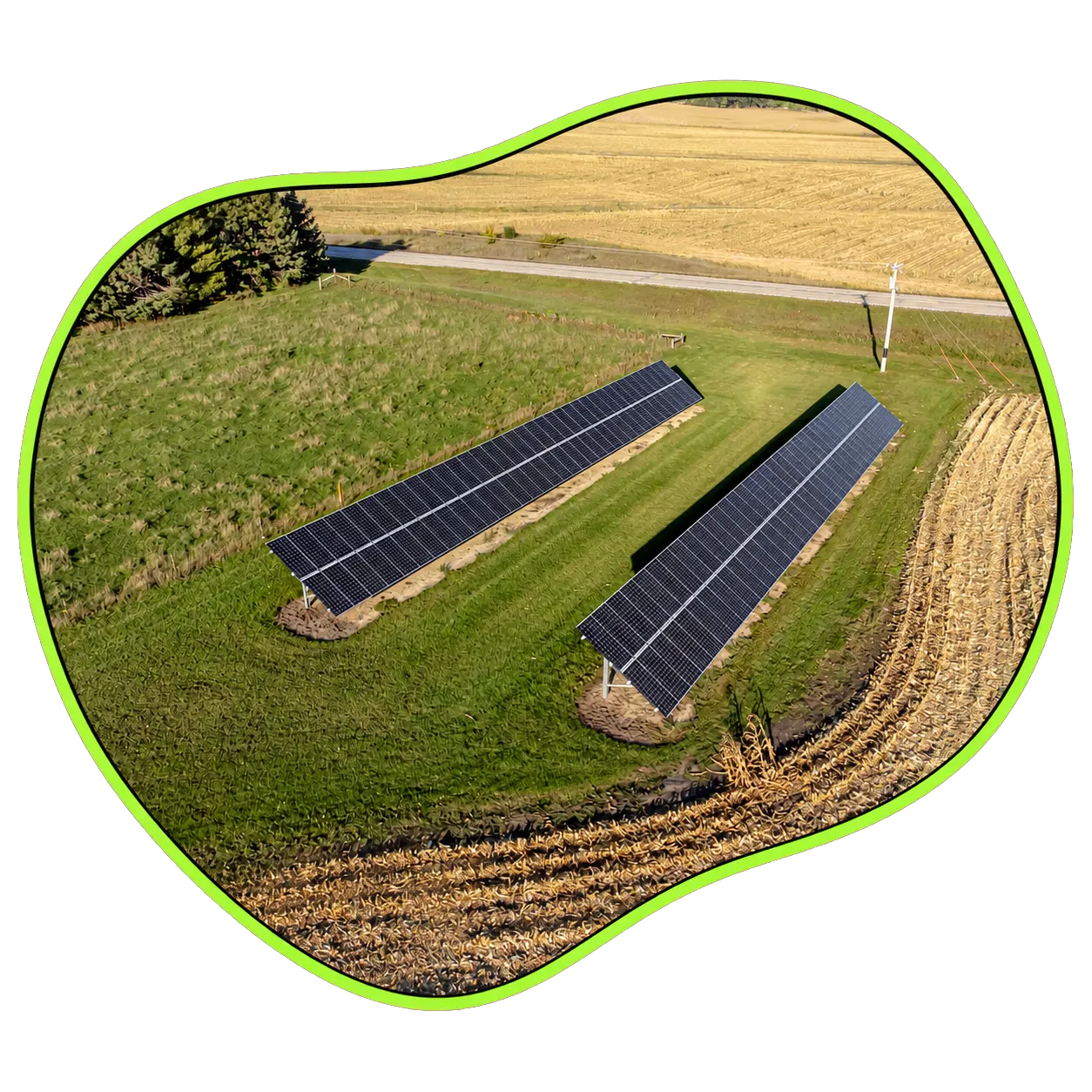 Two rows of solar panels installed on green grass near harvested fields and a road.