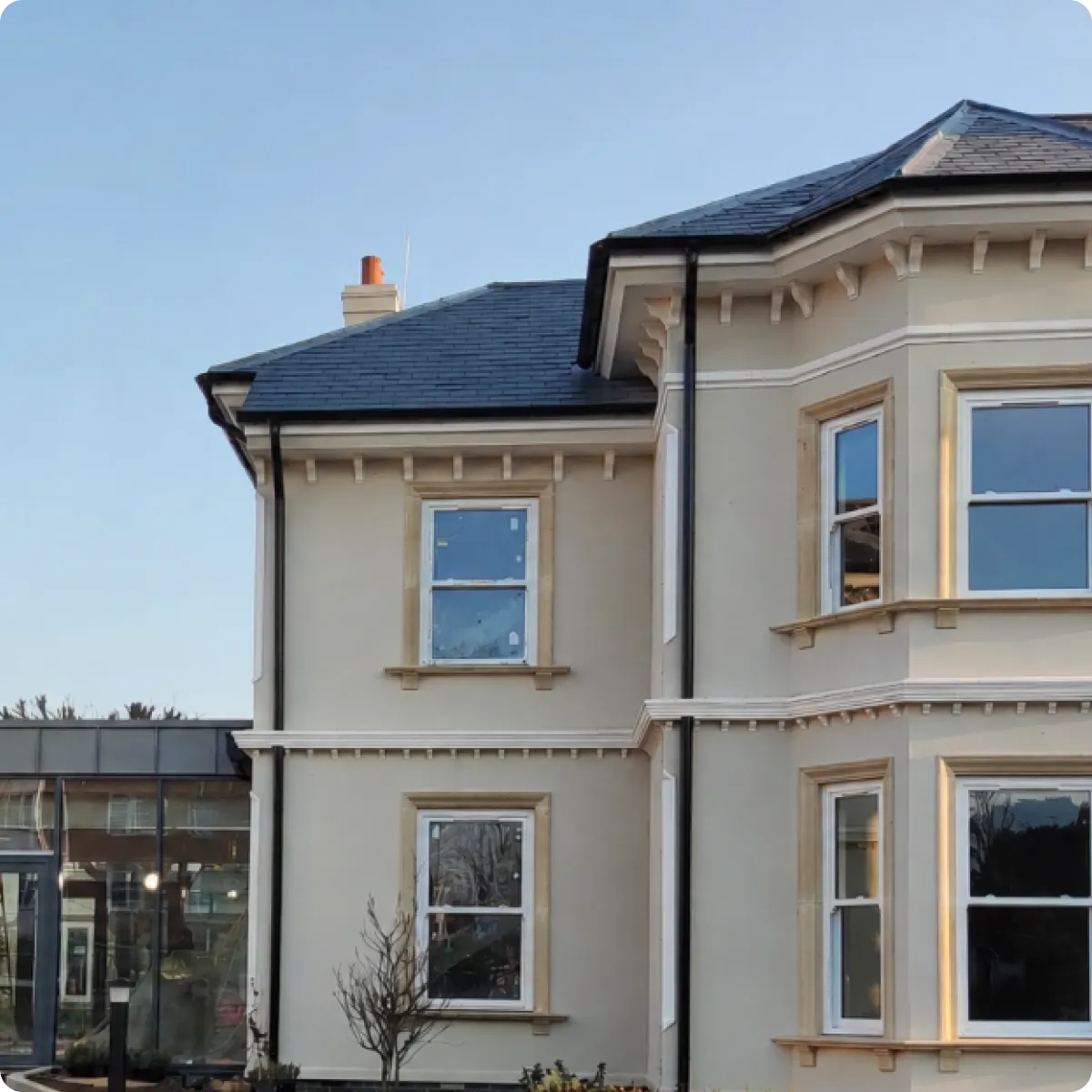 Side view of a beige two-story house with bay windows and a slate roof against a clear sky.