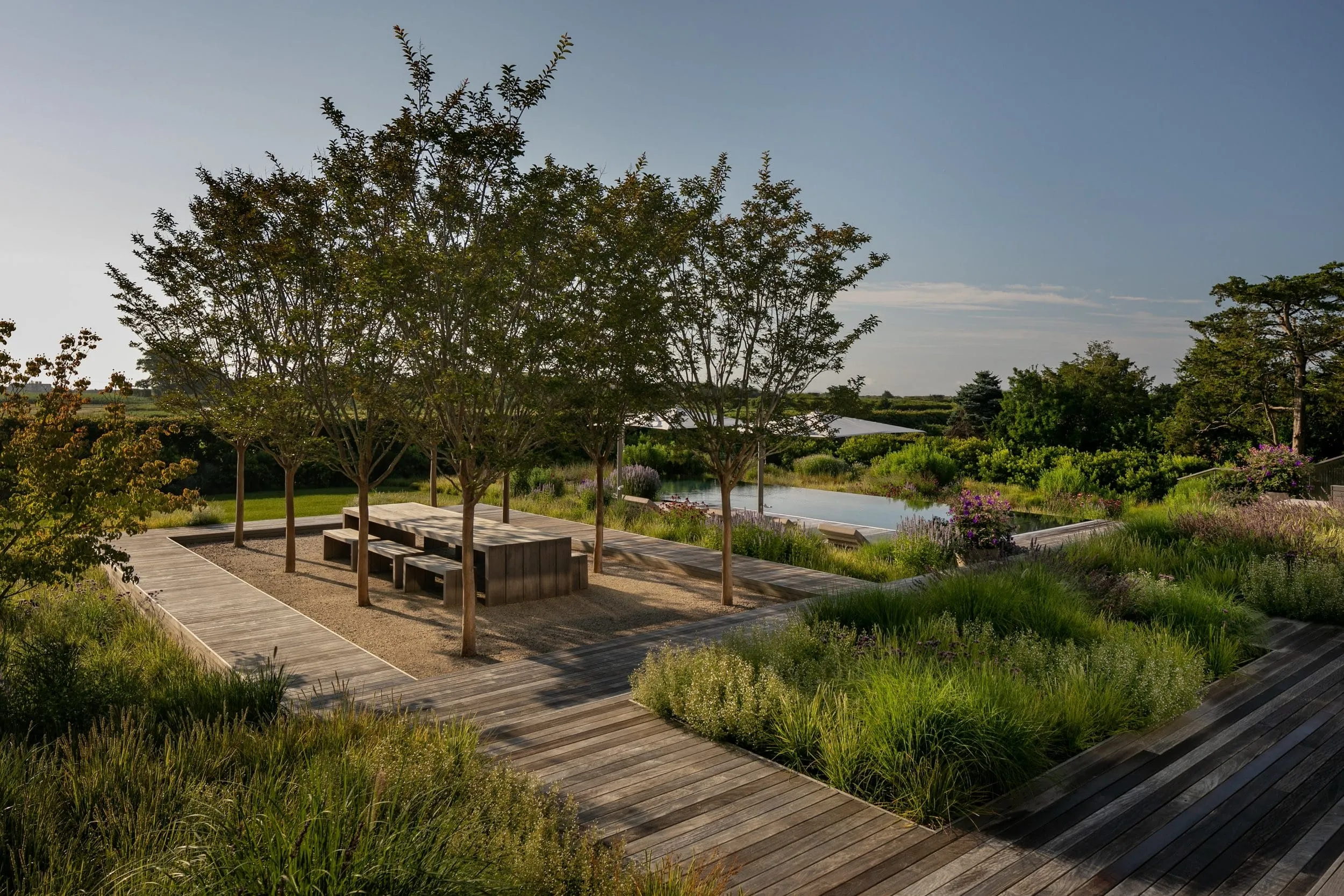 Outdoor wooden picnic table with benches under small trees surrounded by a gravel area and wooden pathways, with lush green plants and a reflective pool in the background.