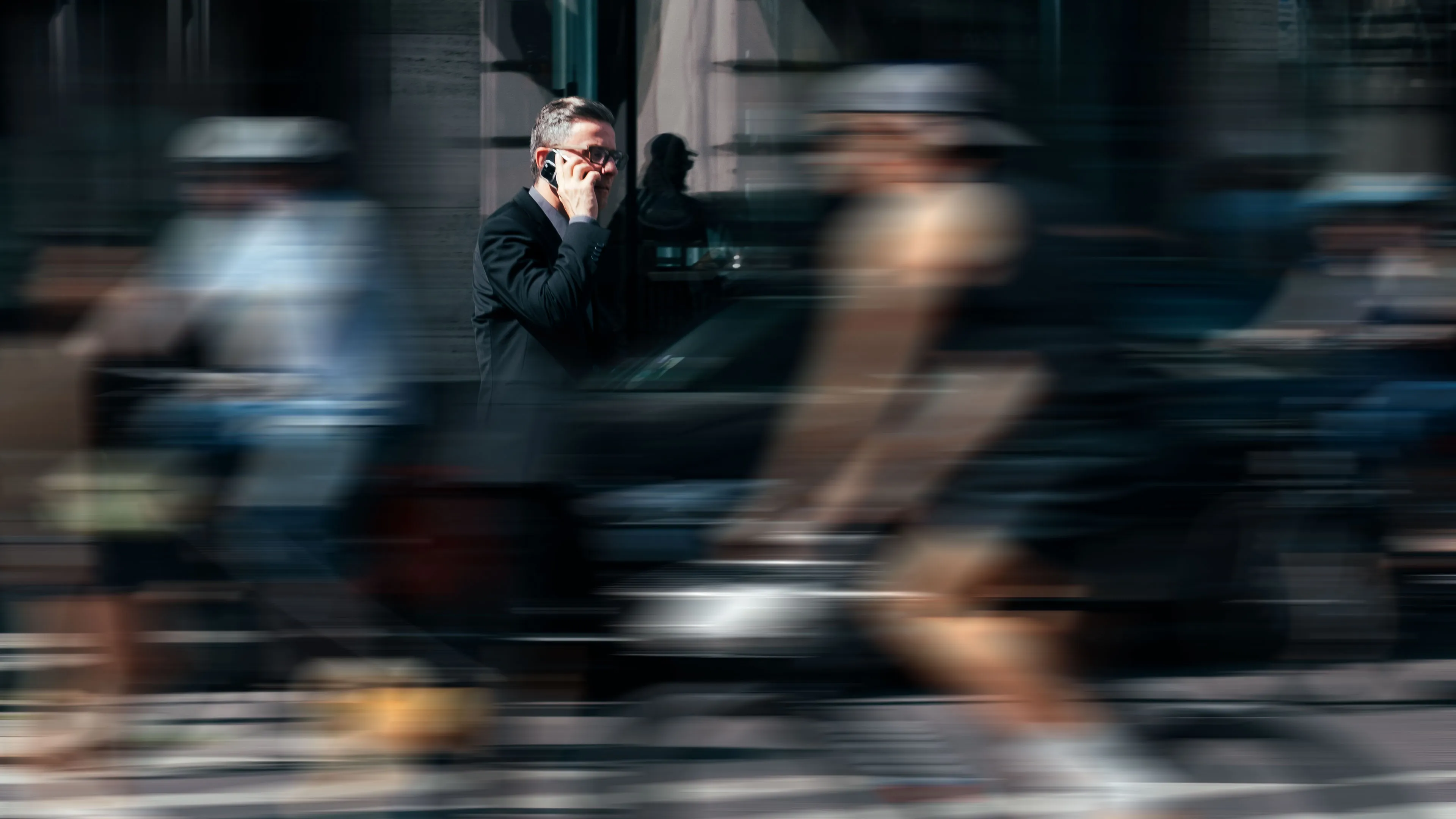 Ein Mann im Anzug telefoniert, während um ihn herum unscharf vorbeifahrende Fahrradfahrer zu sehen sind.