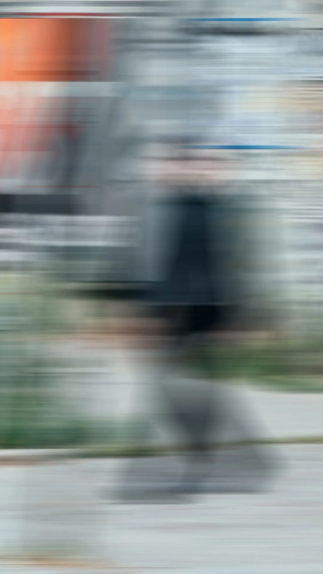 Unscharfes Bild einer Person, die auf einem Bürgersteig geht, mit Gebäuden und Pflanzungen im Hintergrund.