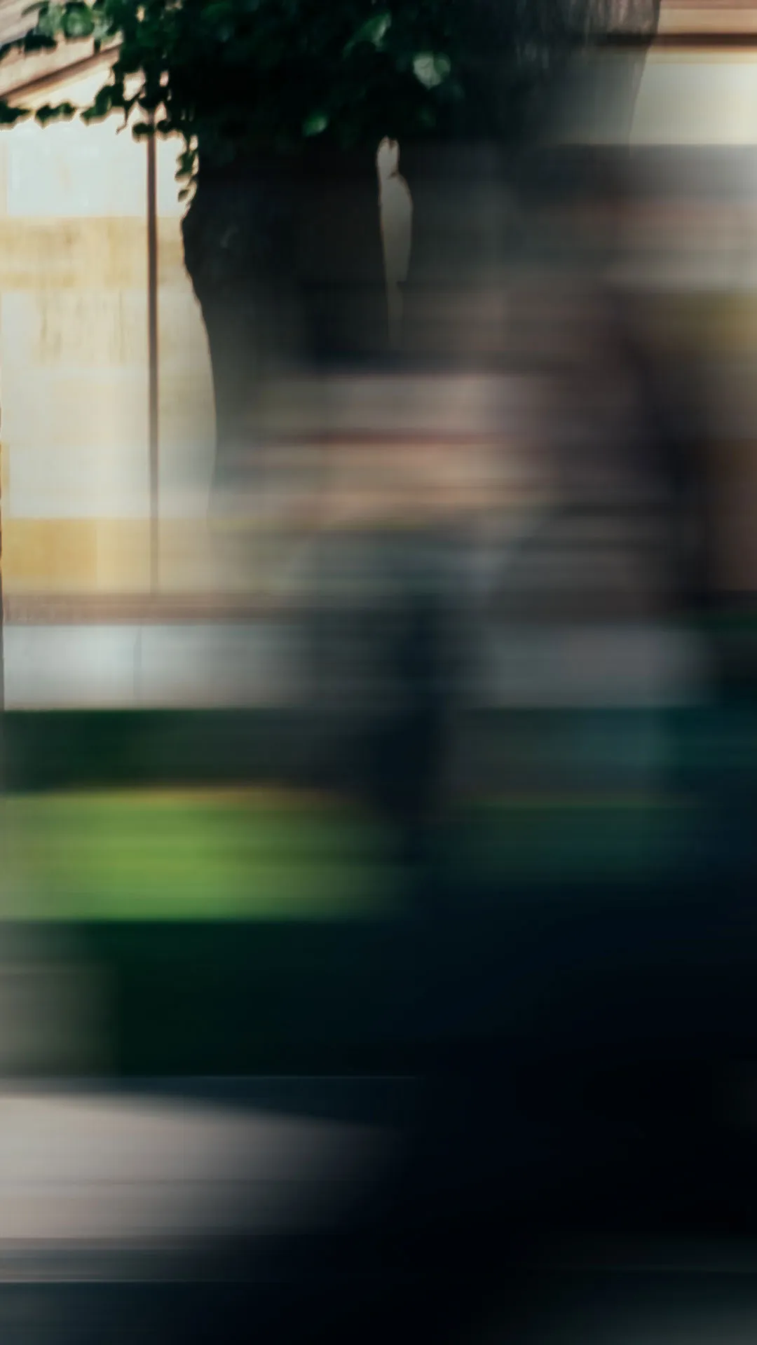 Verschwommene Szene mit vorbeigehender Person vor einem Baum und einer Mauer.