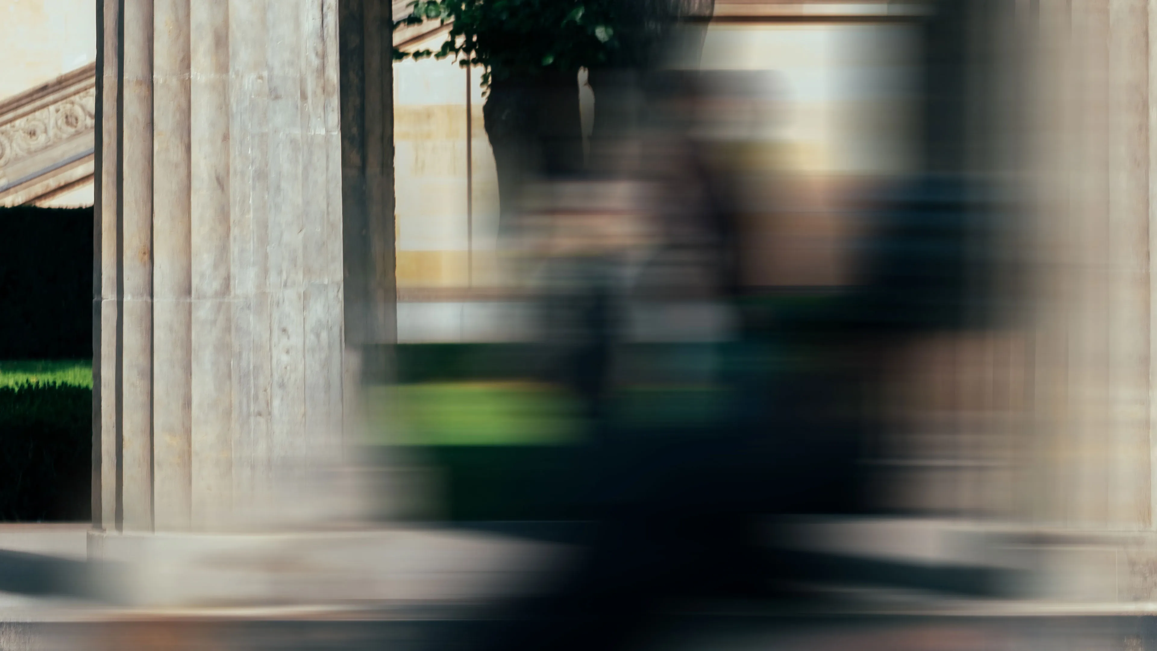 Unschärfe einer Person, die vor einem Gebäude mit Säulen und grünem Garten vorbeigeht.