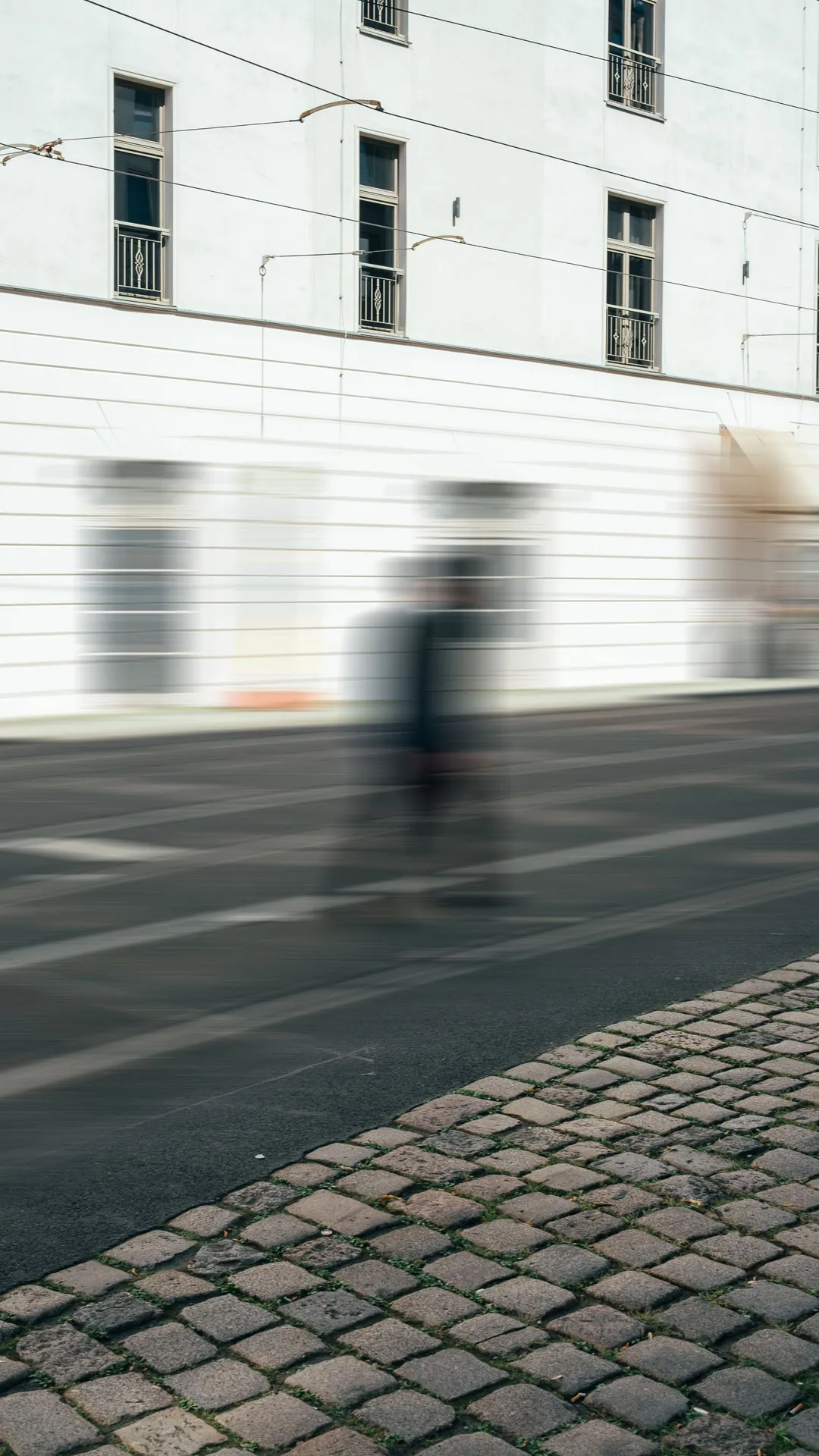 Zwei unscharfe, sich bewegende Personen gehen auf einer Straße vor einem weißen Gebäude mit Fenstern.
