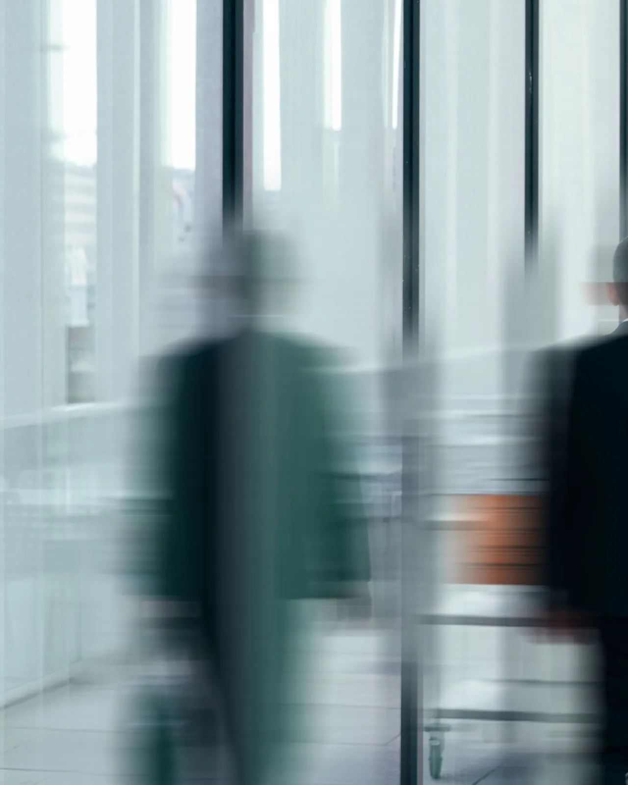 Verschwommene Darstellung von zwei Personen in Anzügen, die sich in einem modernen Gebäude mit großen Glasfenstern bewegen.