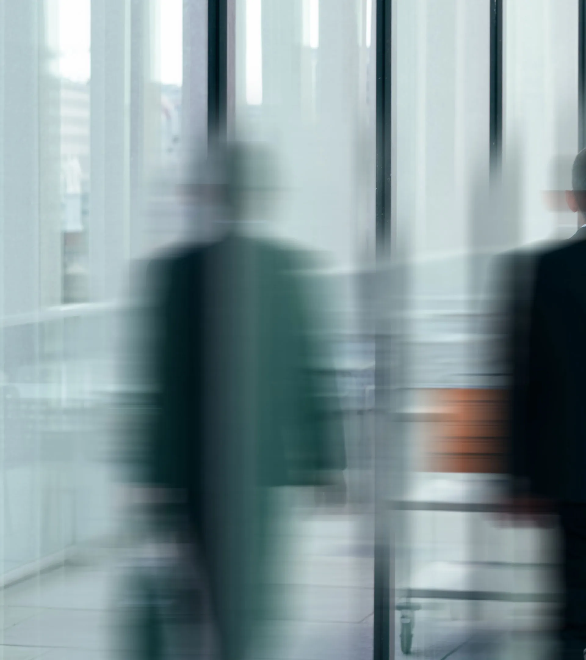Verschwommene Aufnahme von zwei Personen in Geschäftskleidung, die sich in einem modernen Gebäude mit Glaswänden bewegen.