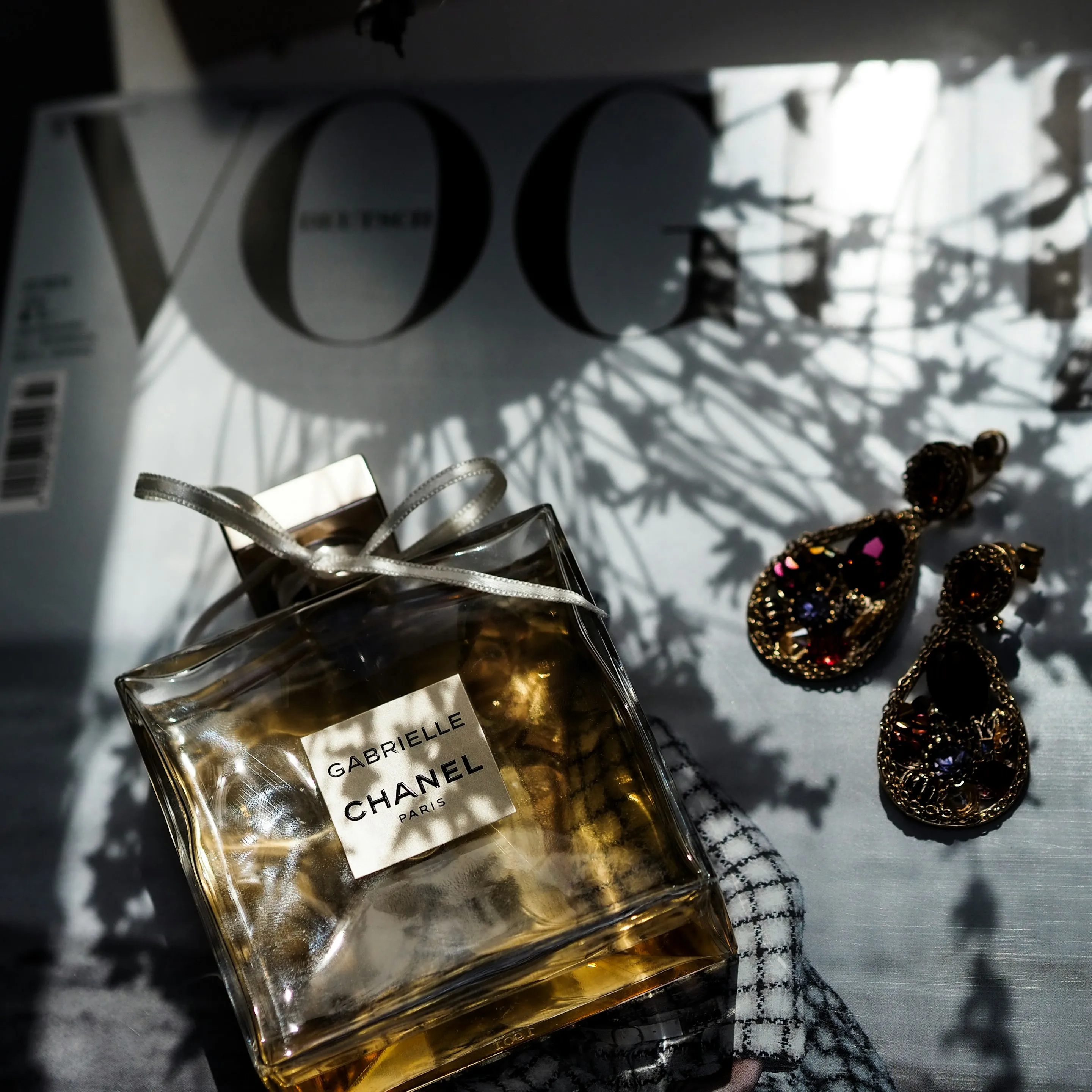 Chanel Gabrielle perfume  beside ornate jeweled earrings on top of Vogue magazine.