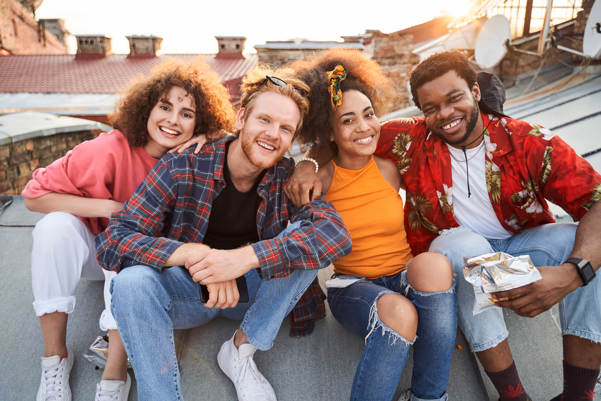 Four young adults sitting close together on a rooftop, smiling and enjoying each other's company.