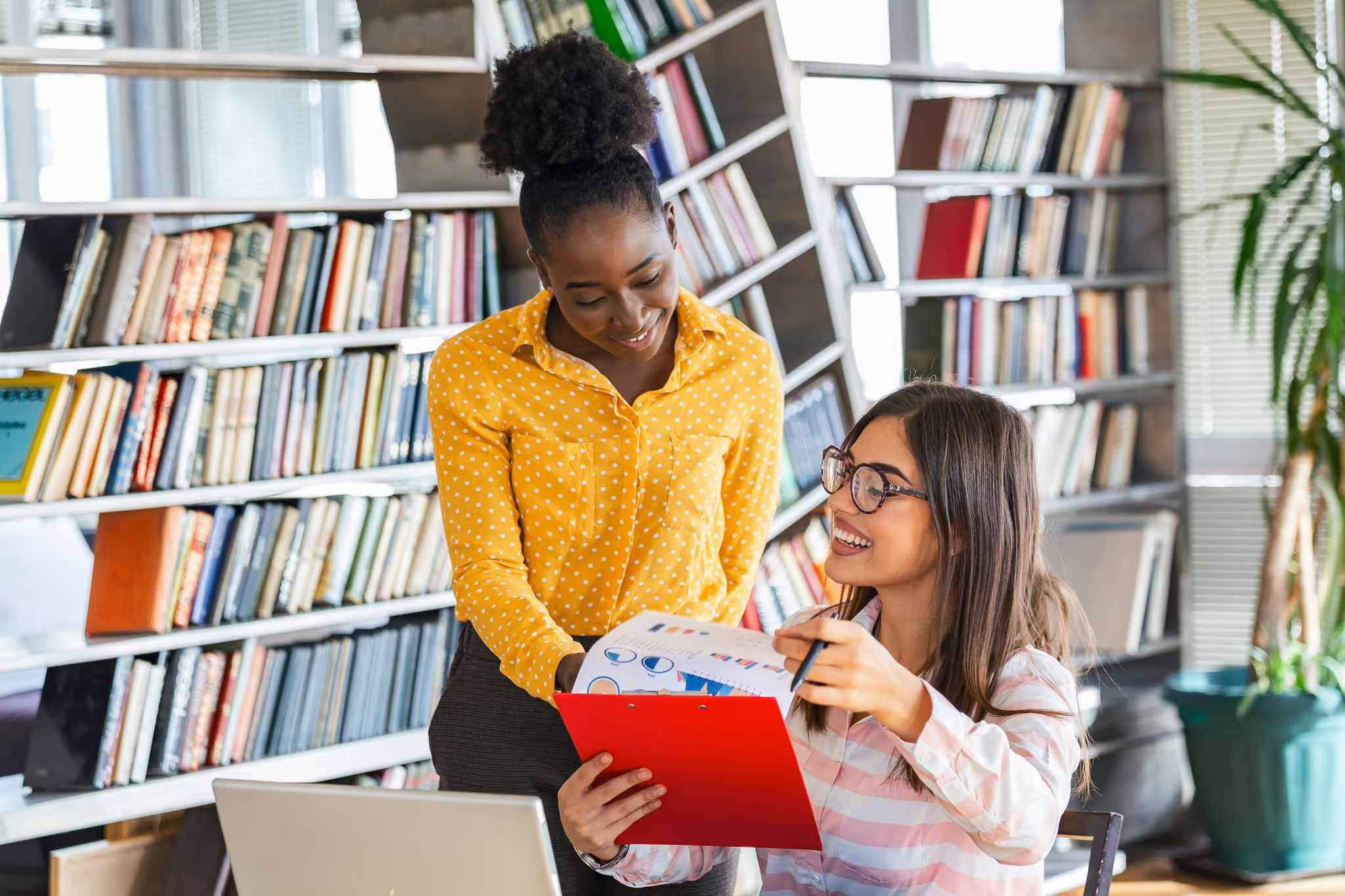 Two women smiling and discussing charts on a clipboard in a library setting with bookshelves in the background.