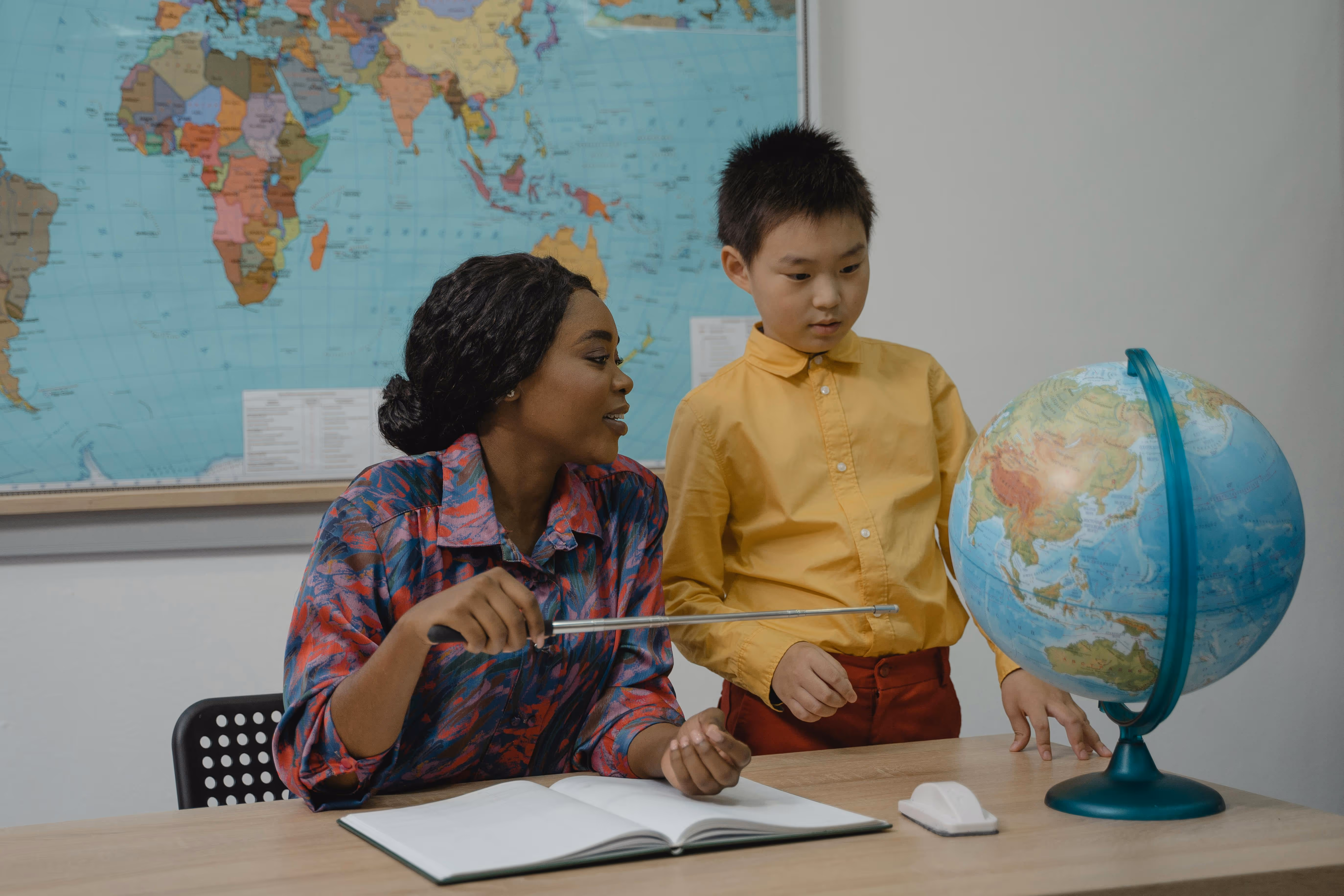 Teacher pointing at a globe with a pointer while explaining to a student who looks at the globe attentively.