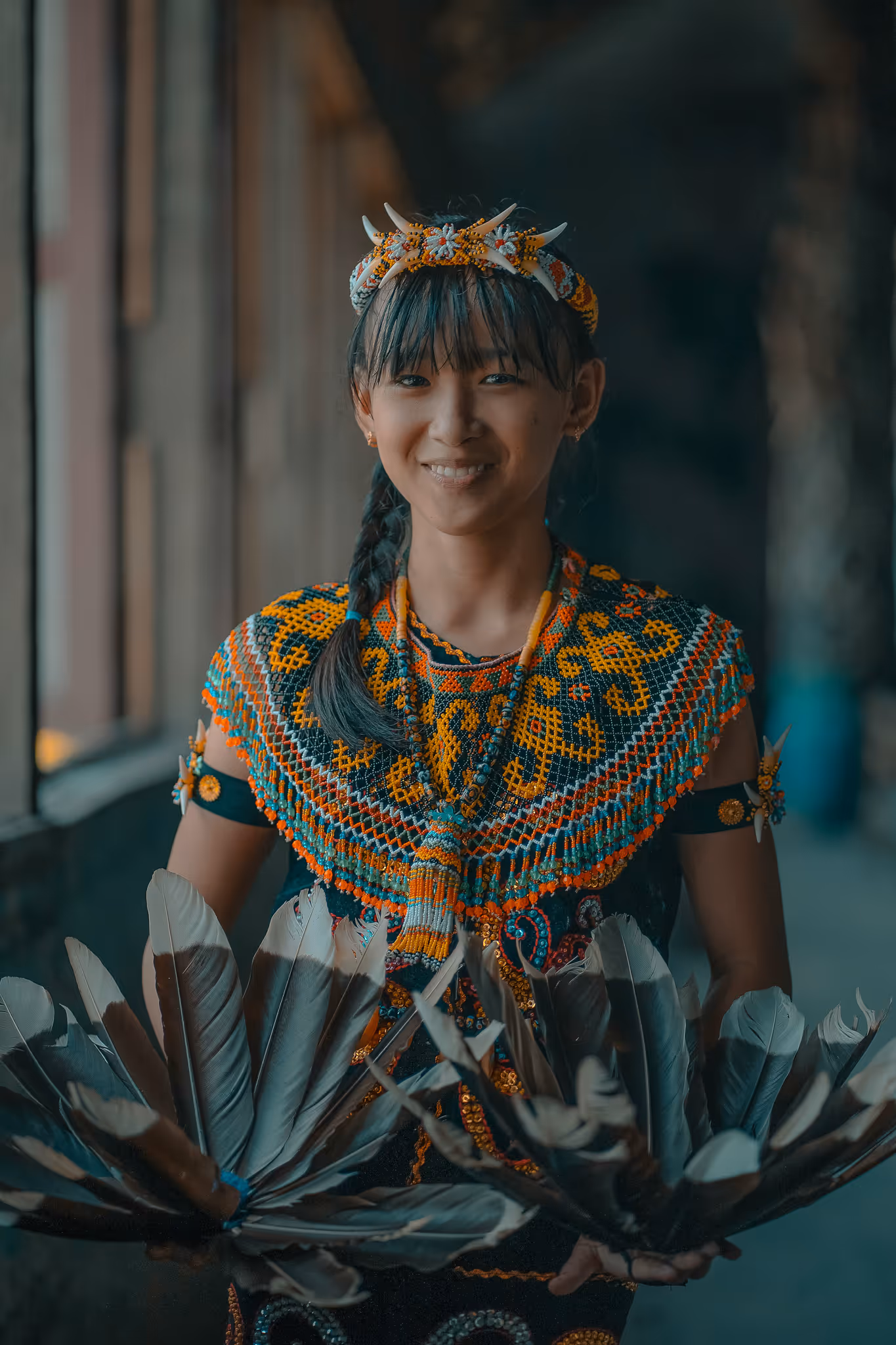 Smiling young woman in colorful traditional beadwork attire holding feathered fans.