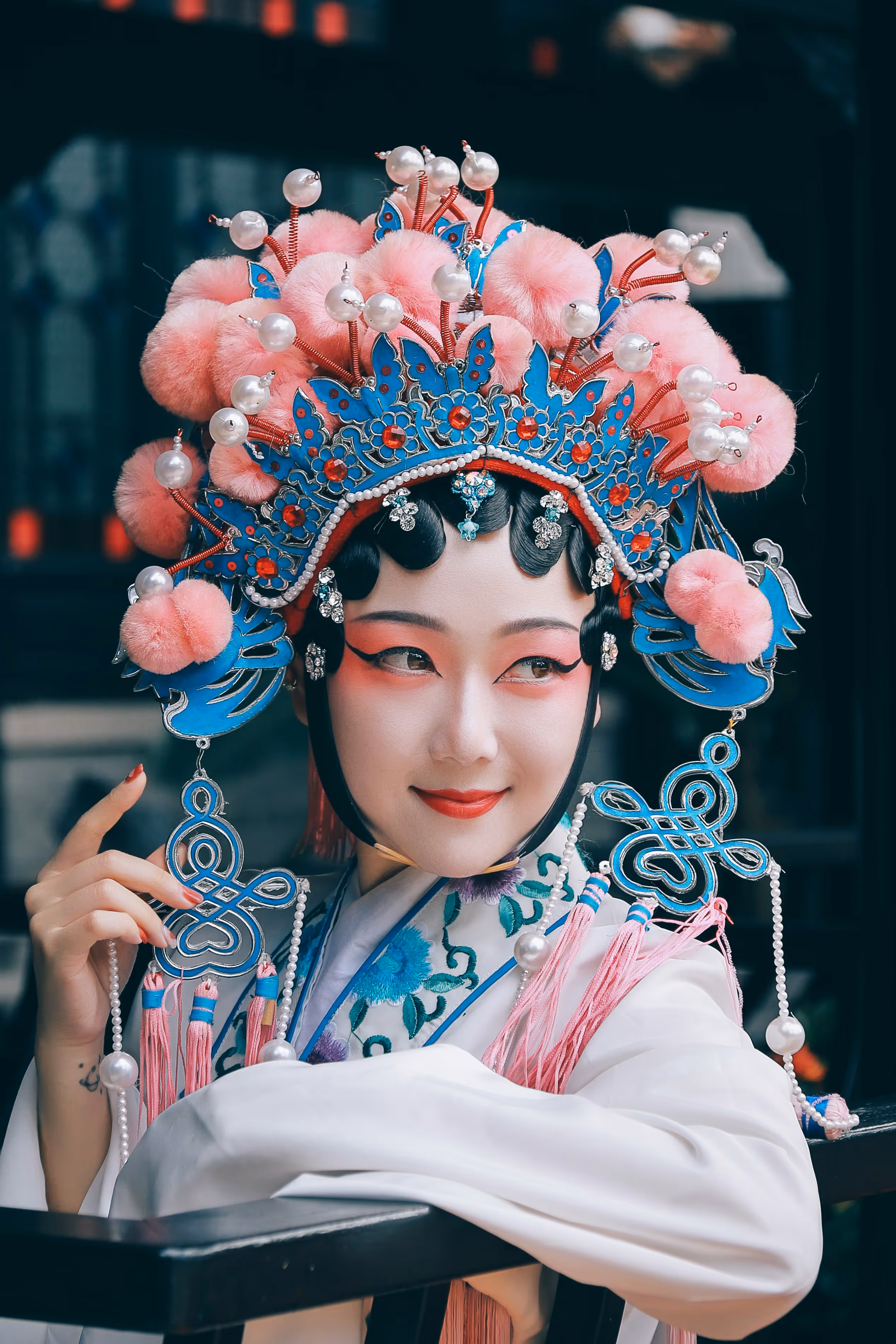 Woman in traditional Chinese opera costume with elaborate blue and pink headdress and embroidered robe smiling and resting arm on chair.