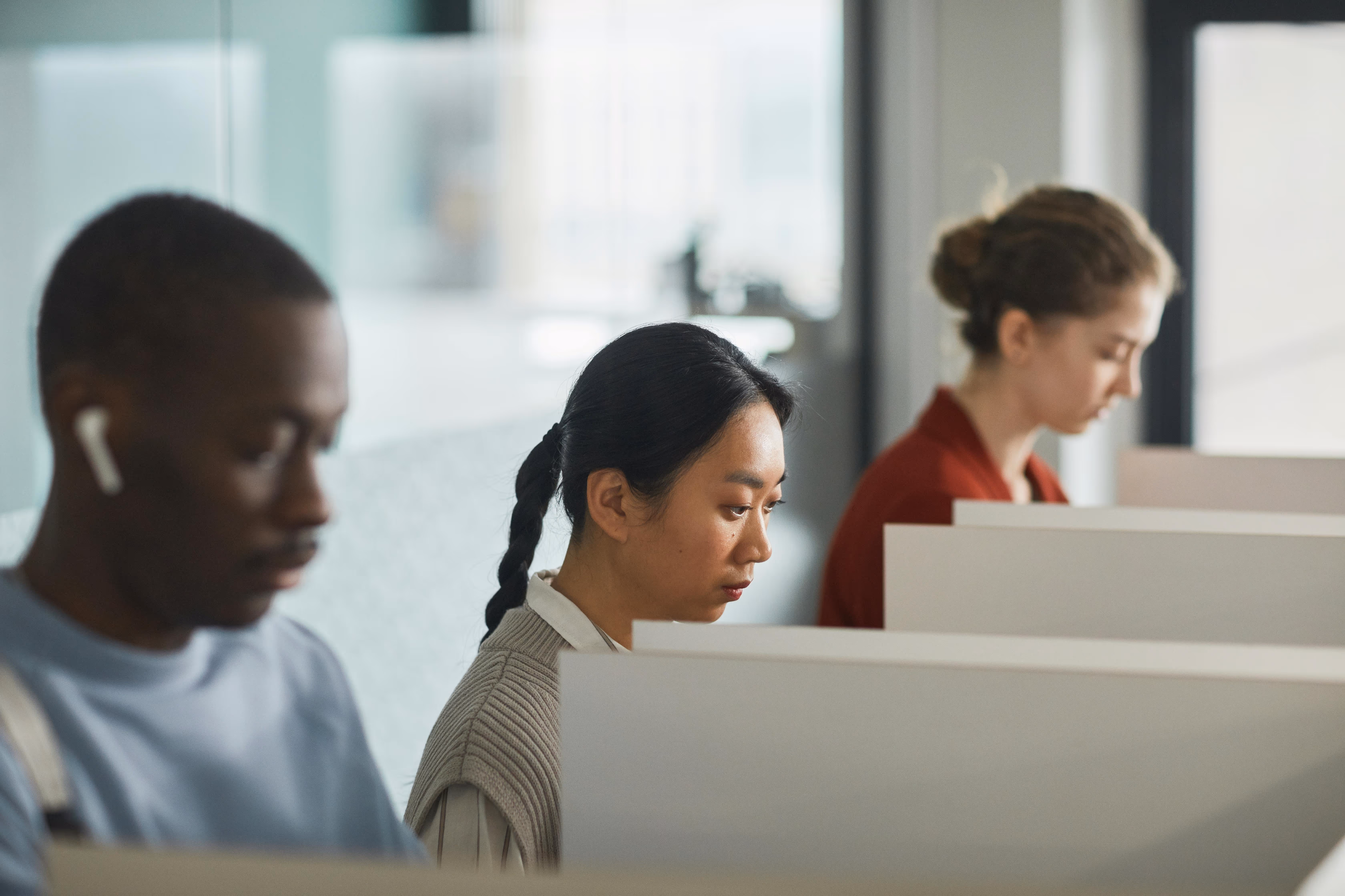 Three people seated in individual work cubicles, focused on their tasks in a bright office.