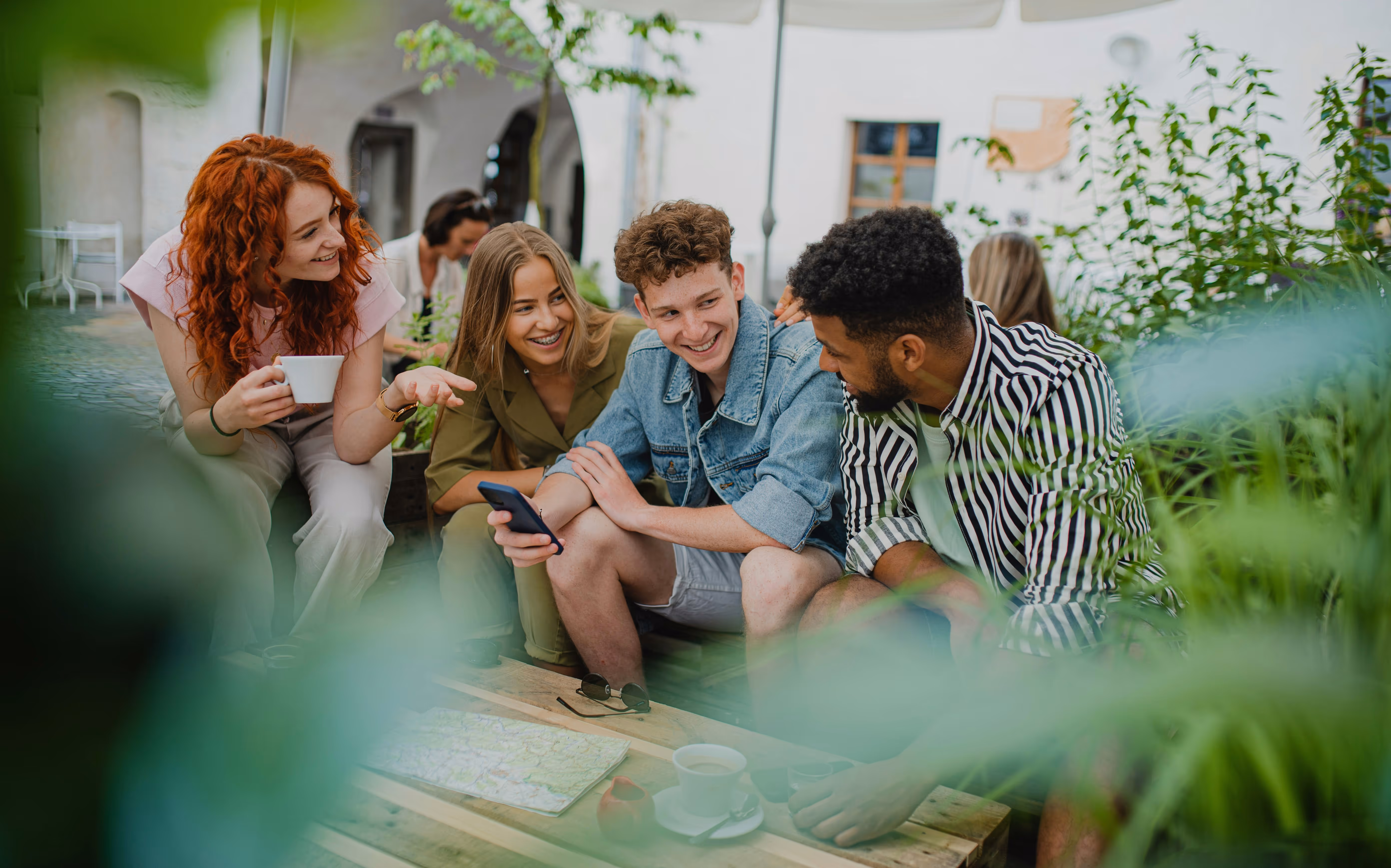Group of four young adults sitting outdoors chatting and smiling, one holding a phone and another holding a coffee cup.