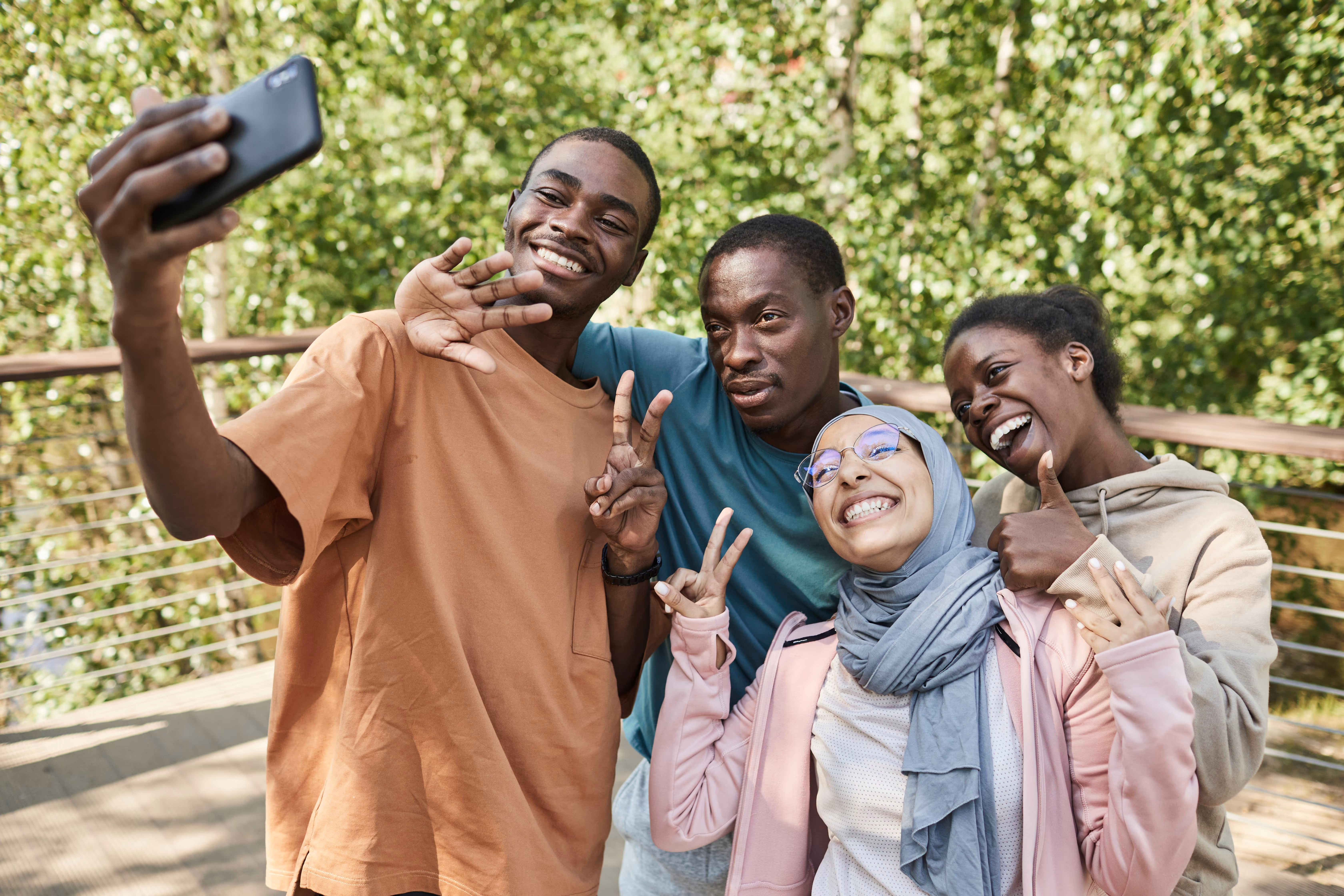 Four friends outdoors taking a selfie with one smiling and waving, two showing peace signs, and one giving a thumbs-up.