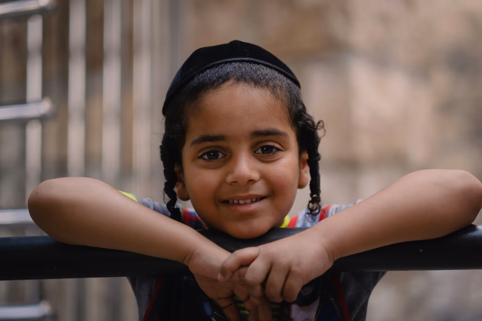 Smiling young boy with curly side locks wearing a black kippah, leaning on a black railing.