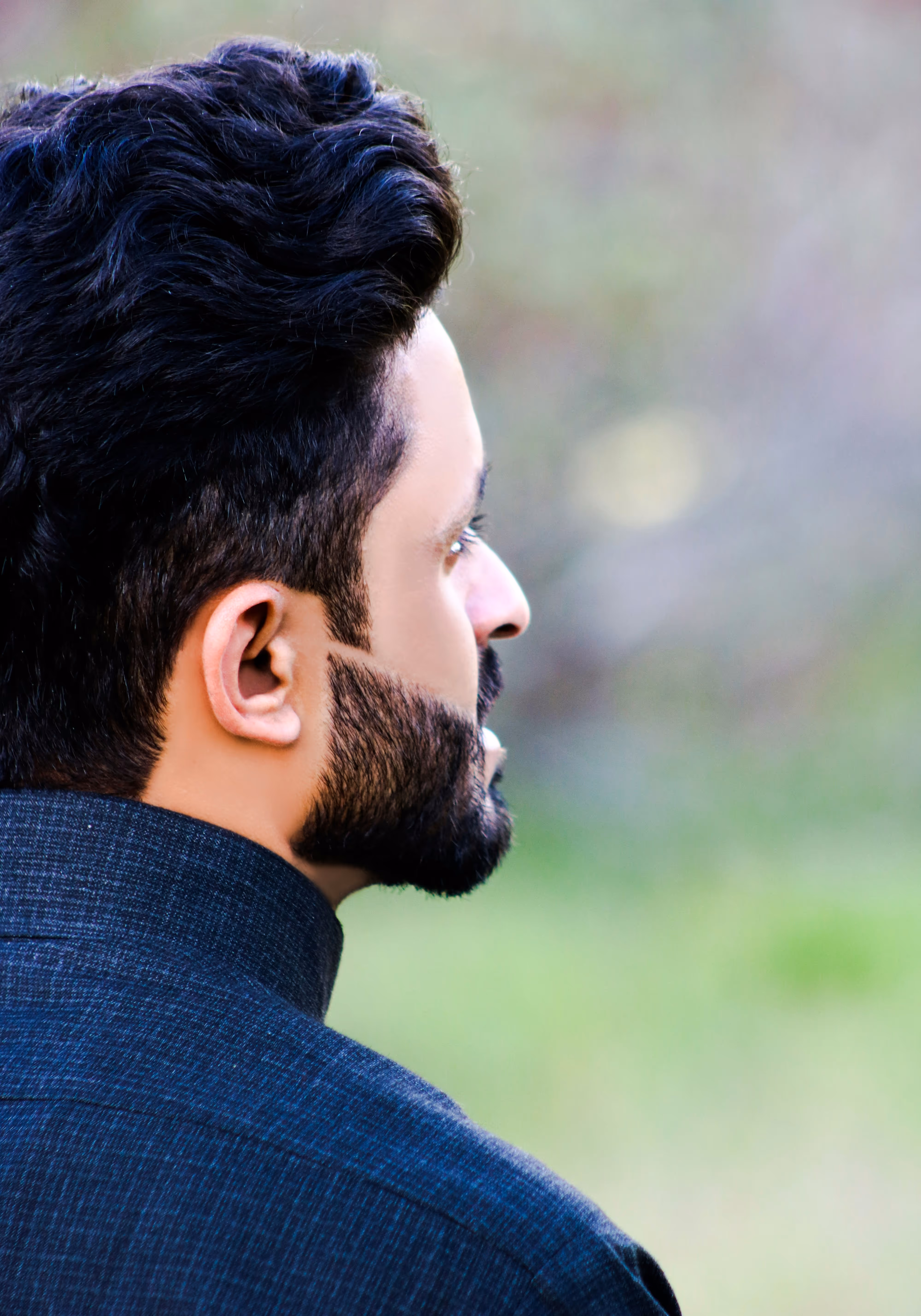 Side profile of a man with styled dark hair and a trimmed beard wearing a dark checkered shirt.