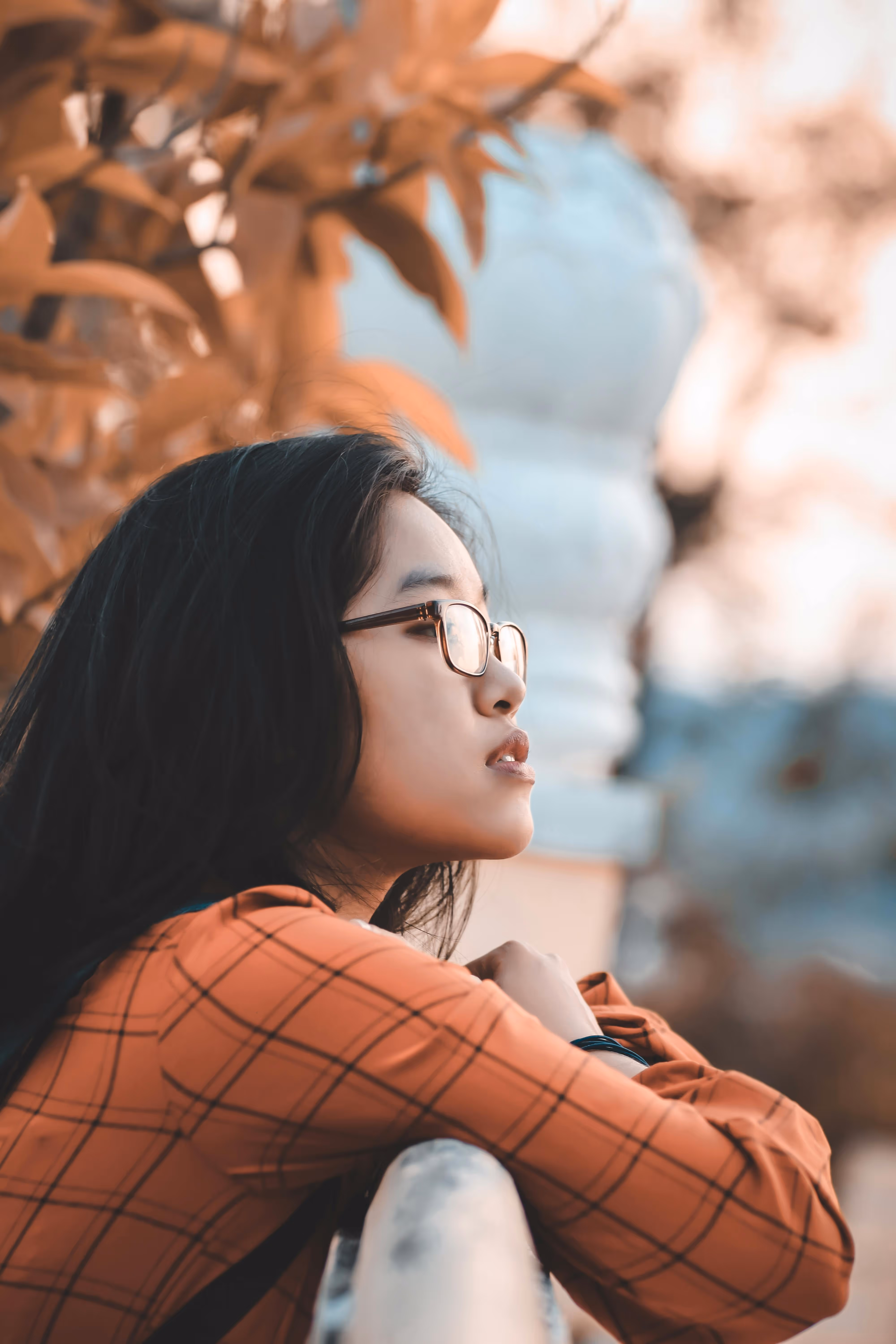Young woman with glasses leaning on a railing, looking thoughtfully into the distance with autumn leaves in the background.