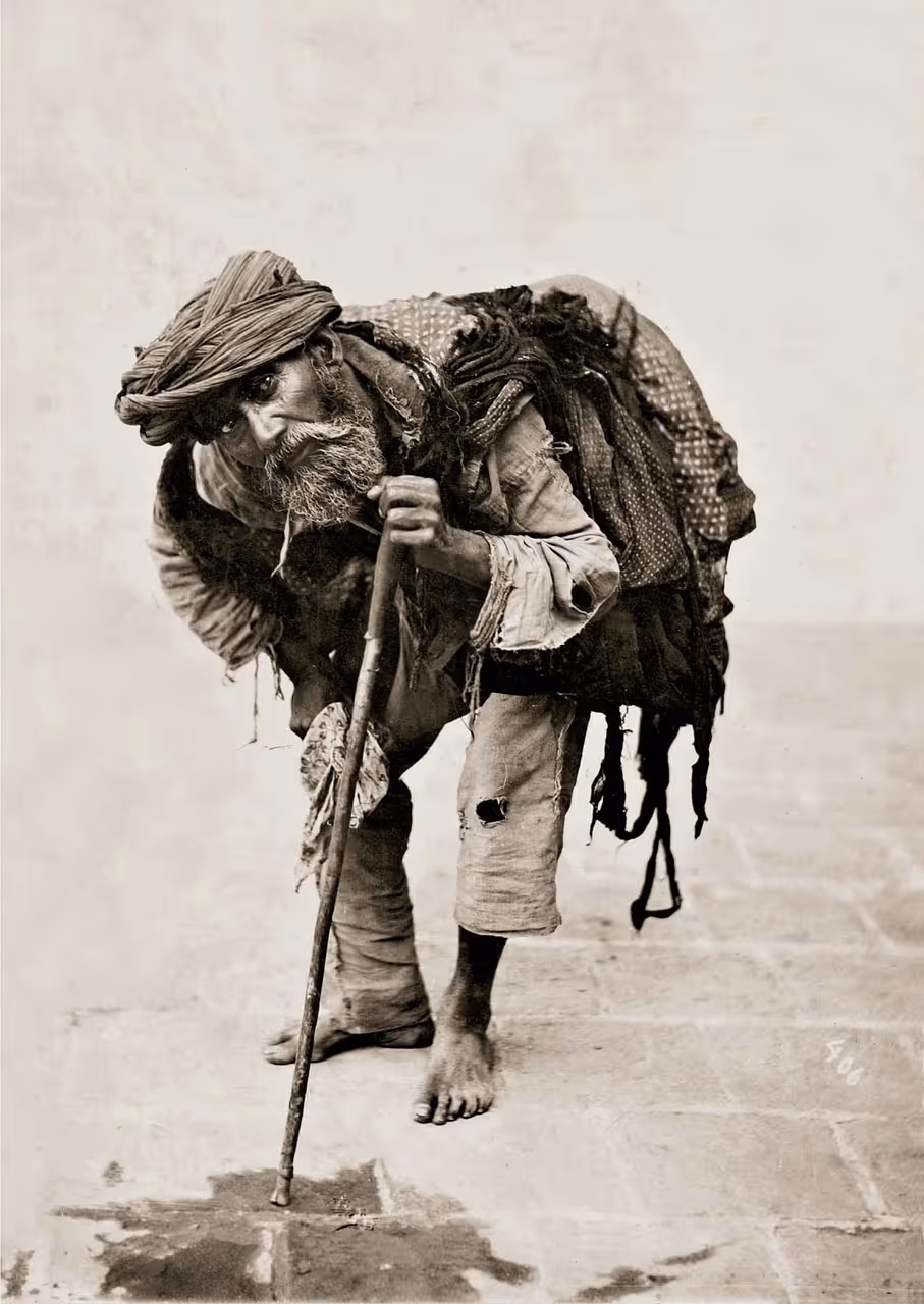Elderly barefoot man with a turban, leaning on a wooden stick and carrying a bundle on his back.
