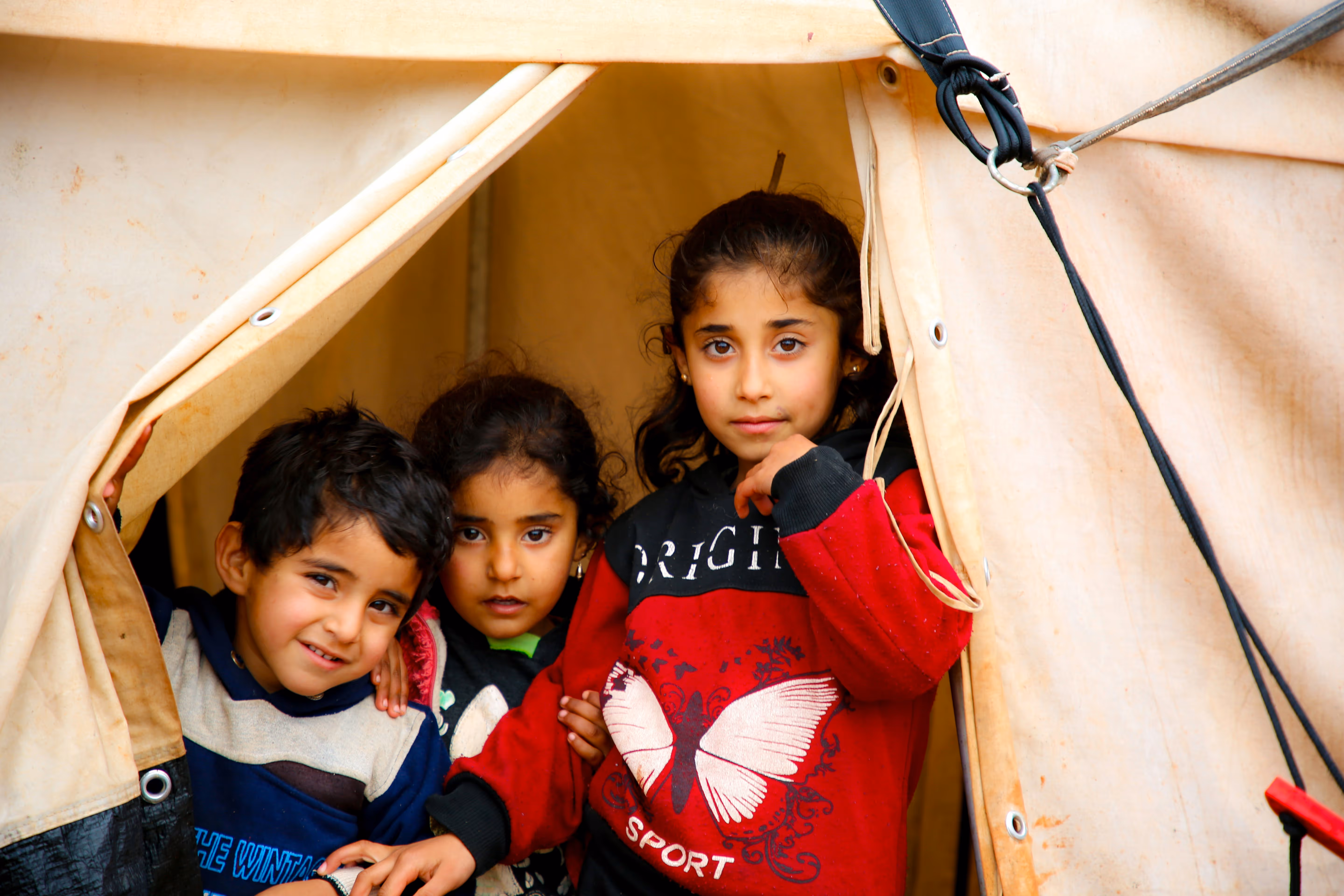 Three children looking out from the opening of a beige tent.