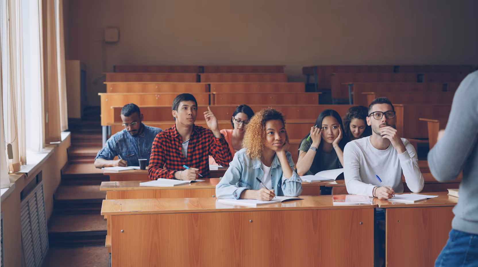 College students sitting in a lecture hall with wooden desks, attentively listening to a professor.