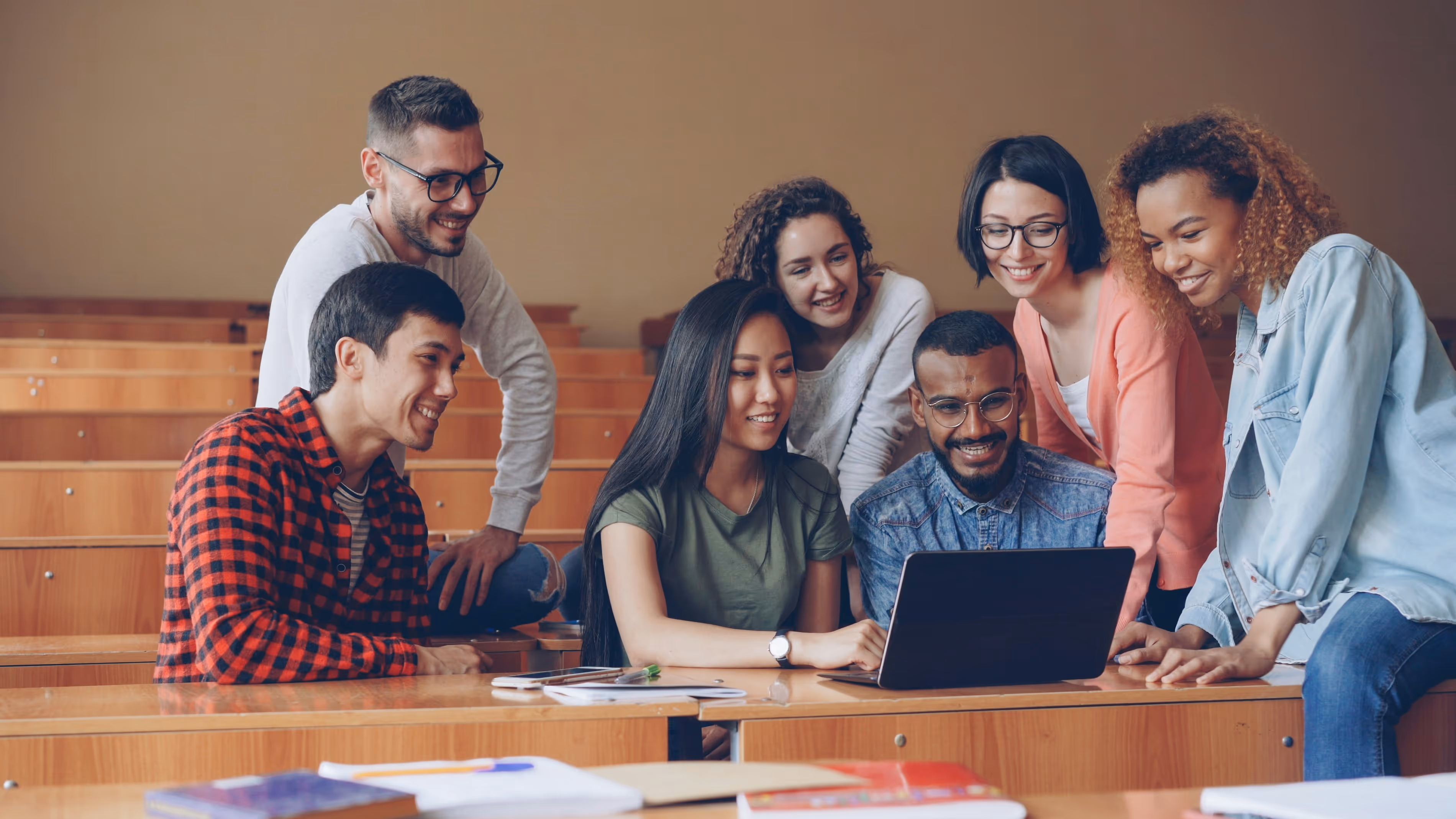 A diverse group of seven young adults smiling and gathered around a laptop in a classroom with wooden desks.