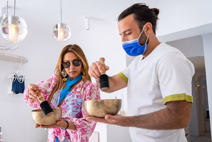 A man wearing a blue face mask and Escarle Silva in sunglasses use mallets to play brass singing bowls indoors.