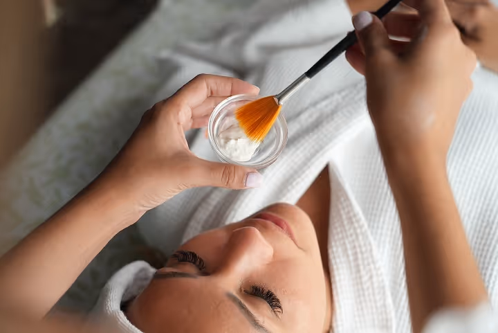 Person holding a bowl of cream and a brush preparing for a facial treatment while lying down wrapped in a white robe.