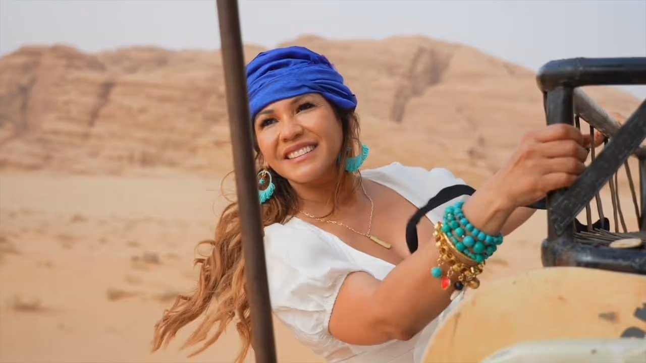 Escarle Silva smiling woman wearing a blue headscarf and turquoise jewelry, holding onto a metal bar in a desert landscape.