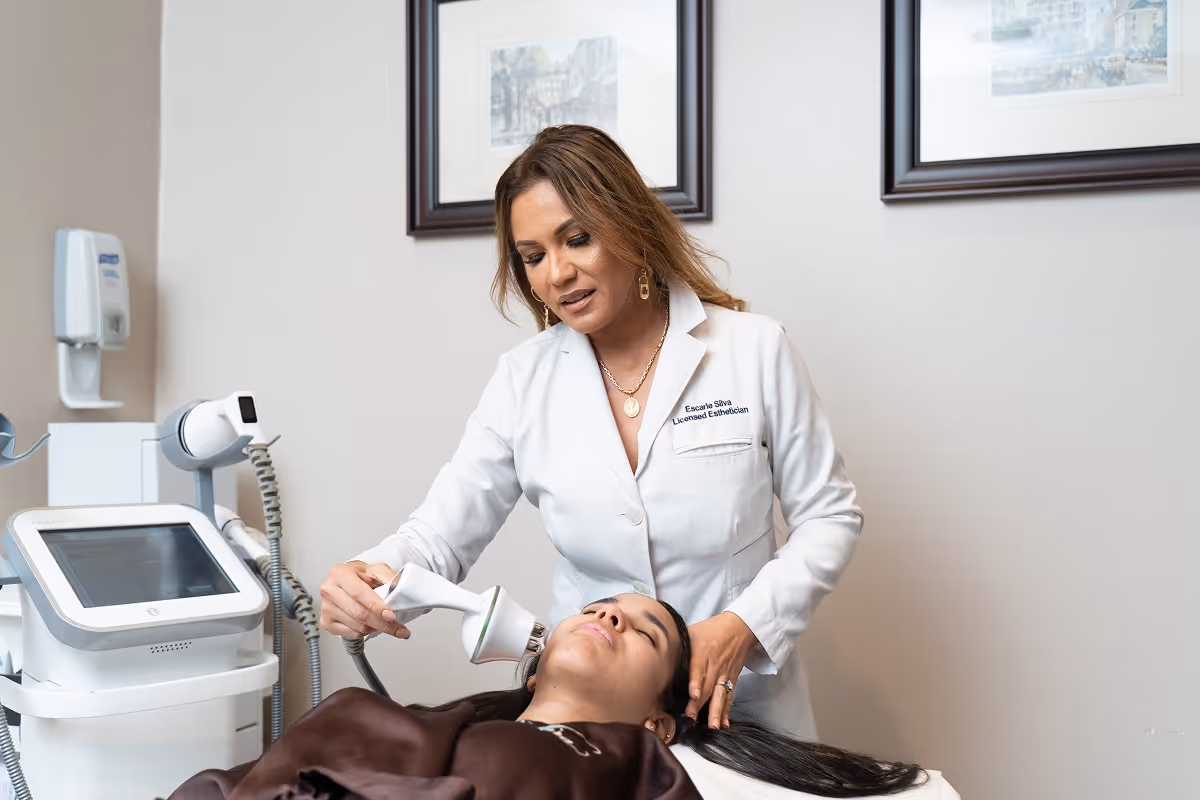 Escarle Silva performing a facial laser treatment using a handheld device on a reclining woman in a clinical setting.