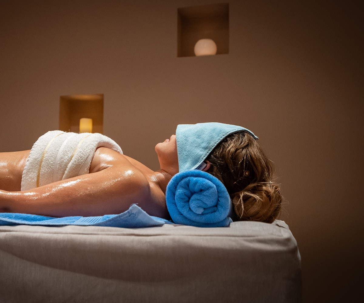 Woman lying face up on spa table with oil on her skin, a towel over her eyes, and a rolled blue towel supporting her head.