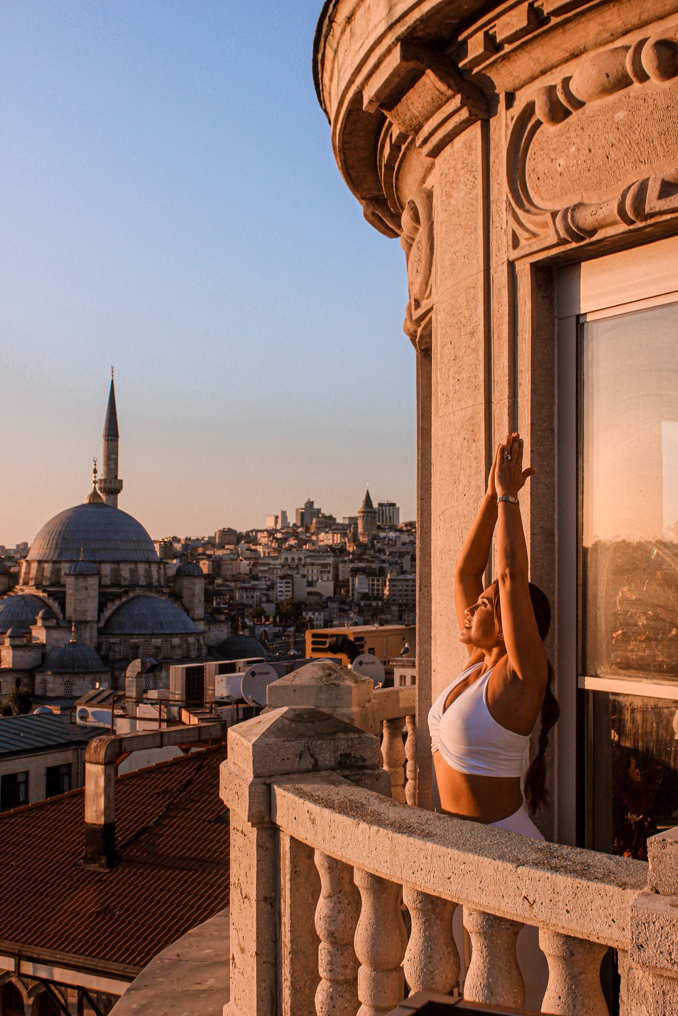 Woman in white workout clothes stretching with arms raised on a stone balcony overlooking a city with mosque domes at sunset.
