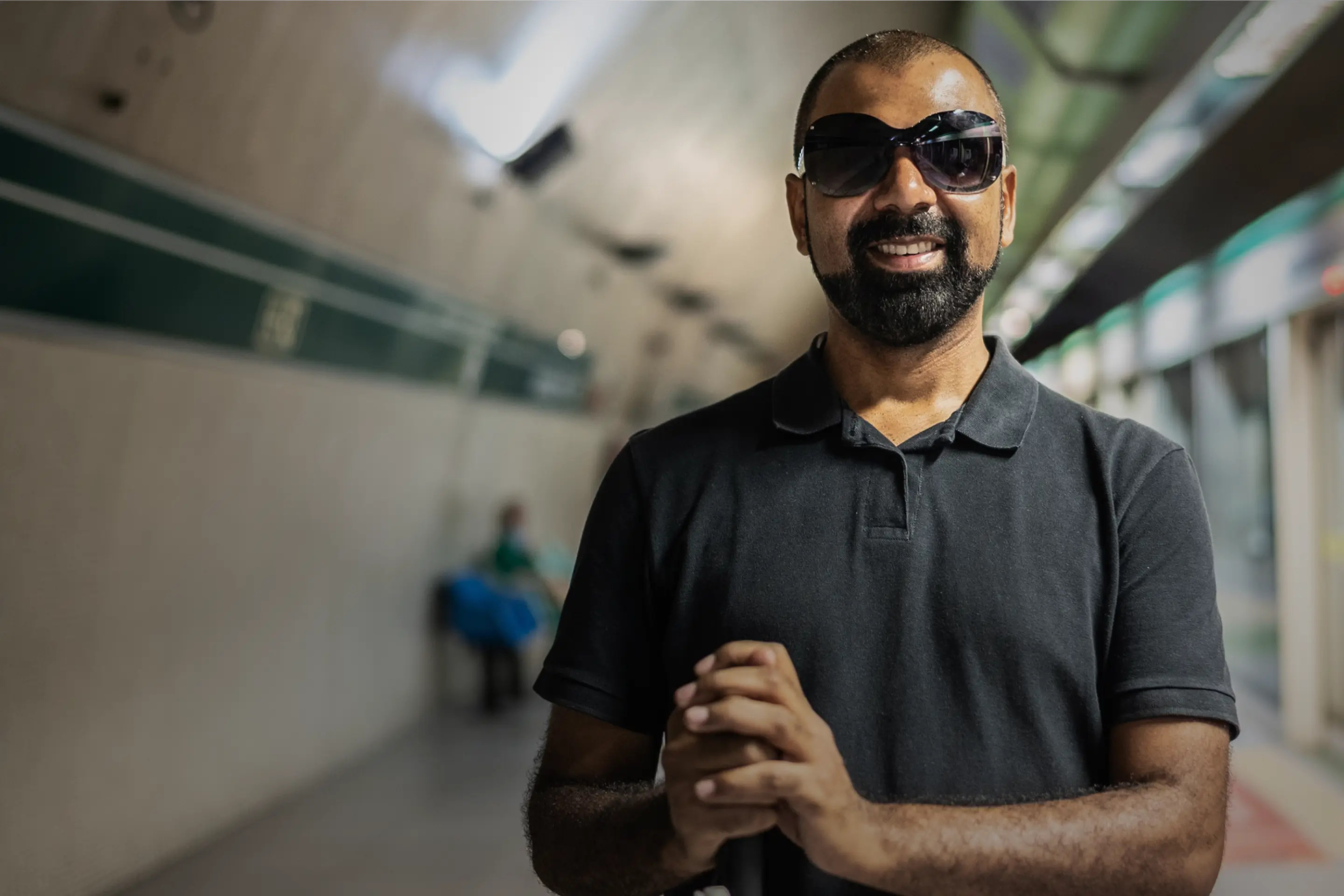 Man standing in a subway smiling at the camera with glasses holding a cane to help with vision challenges.