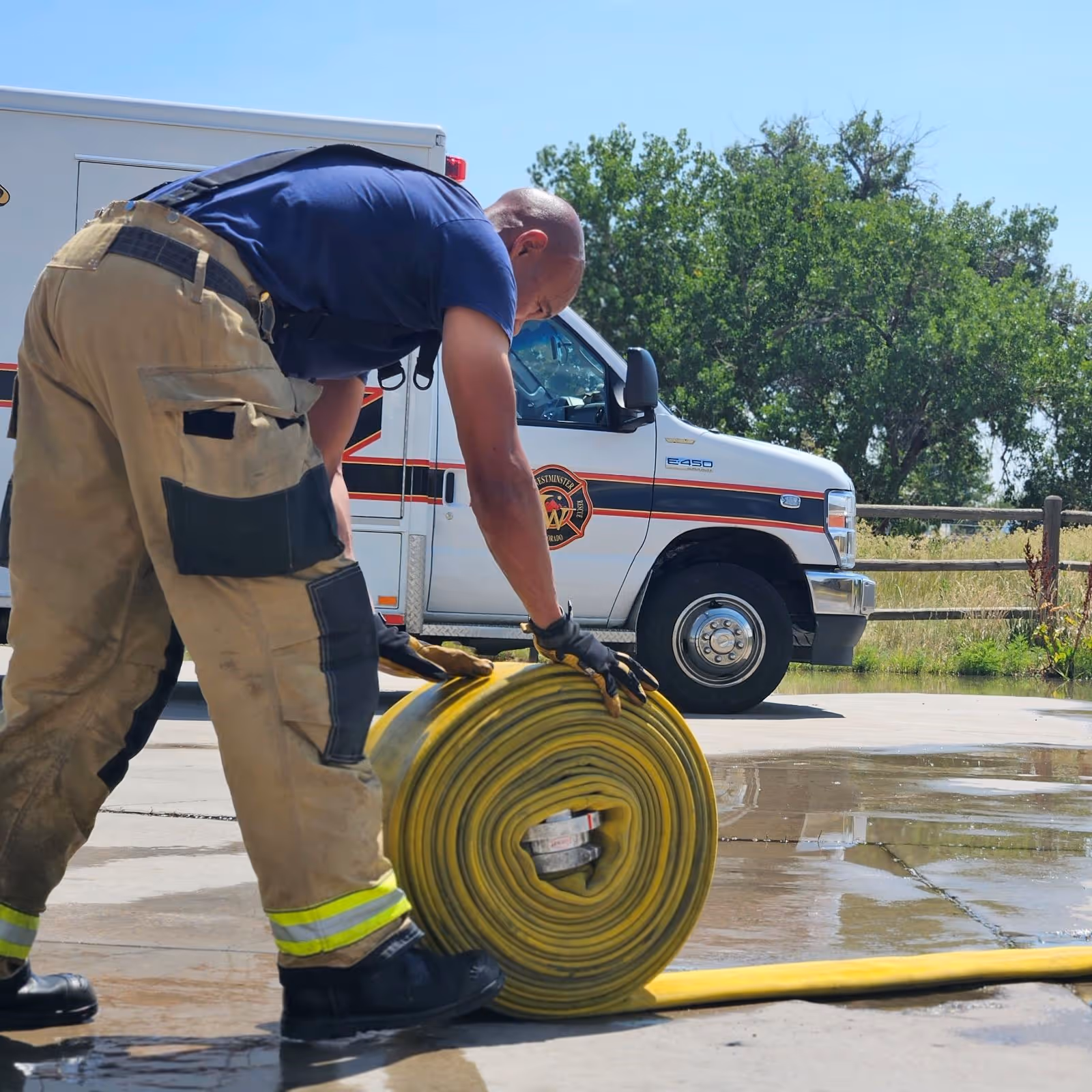  Firefighter rolling up a yellow fire hose near an emergency response vehicle on a sunny day.