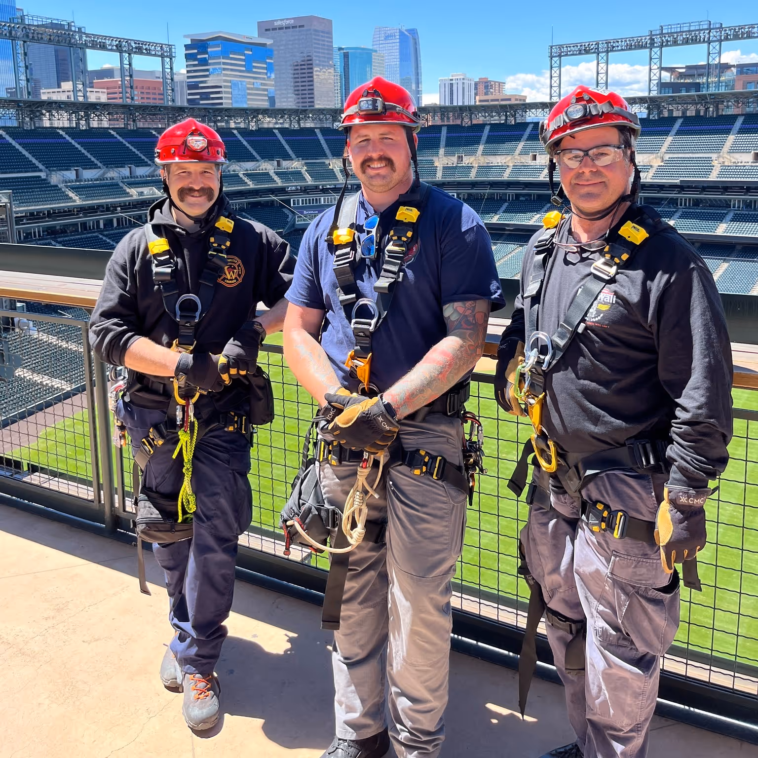 Three firefighters in harnesses and red helmets posing for a photo at a stadium with a city skyline in the background.