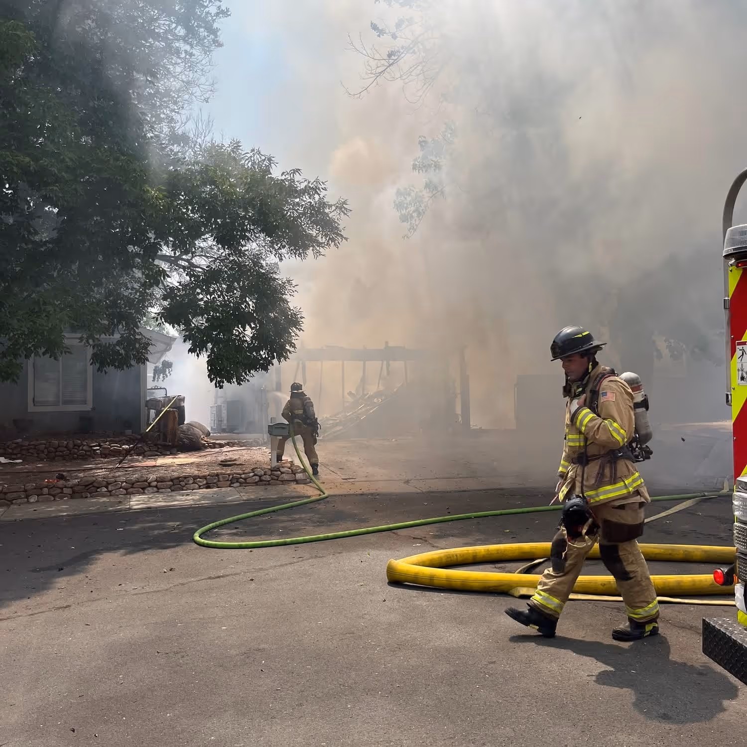 Firefighters responding to a structure fire with heavy smoke, surrounded by trees and hose lines.