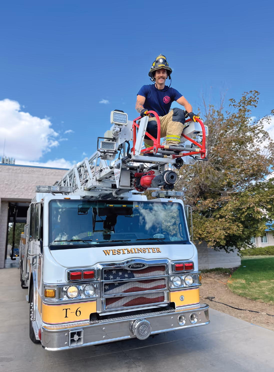 Firefighter sitting on the extended aerial ladder of Westminster Truck 6, smiling under a blue sky.