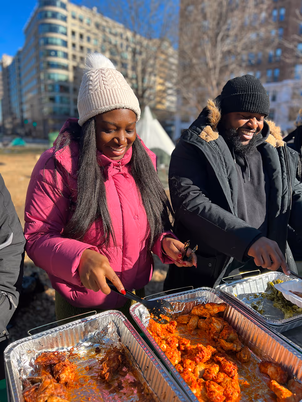 Two volunteers smiling while serving trays of hot food at an outdoor event, wearing winter coats and beanies.