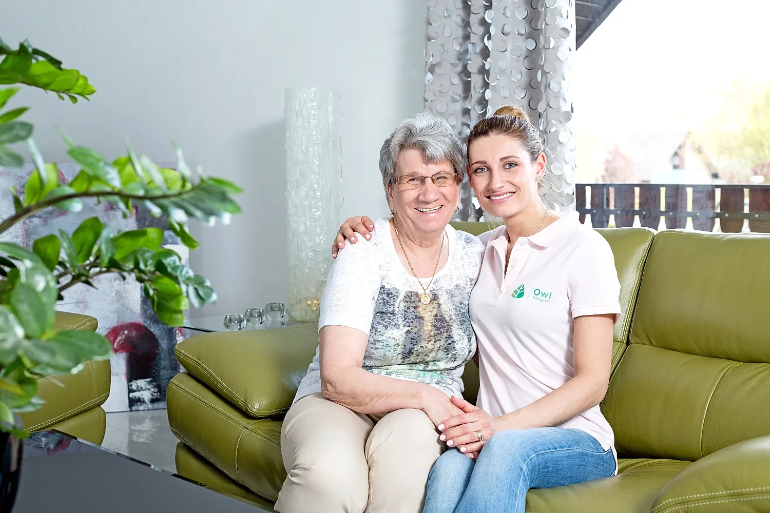 Ältere Frau und junge Pflegekraft sitzen zusammen auf einem grünen Sofa und lächeln.