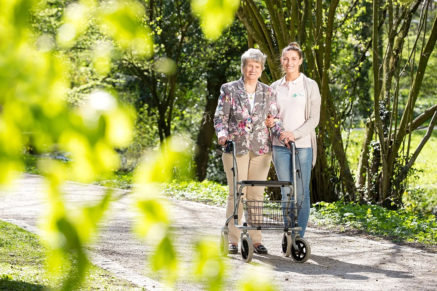 Ältere Frau mit Rollator wird von einer polnischen Pflegekraft bei einem Spaziergang im Park in Paderborn unterstützt.