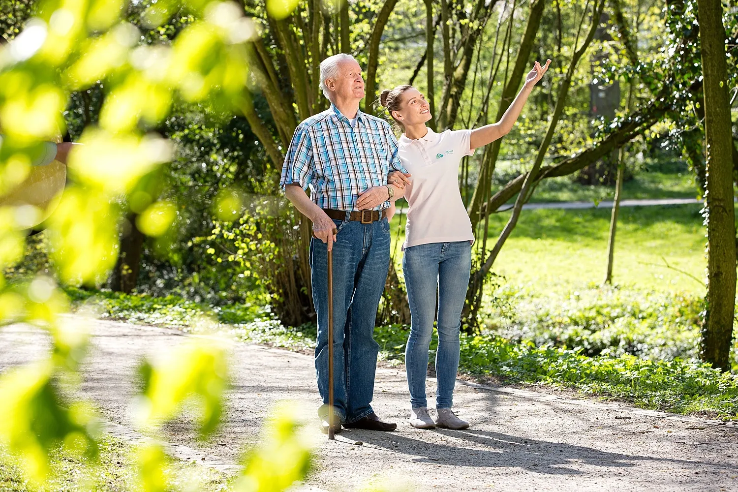 Junge Frau zeigt älterem Mann mit Gehstock etwas in einem sonnigen Park.