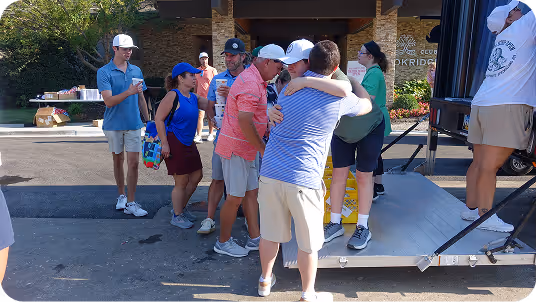 Group of people, including children and adults, gathered outdoors near a stage with some hugging and others standing nearby in casual summer clothing.