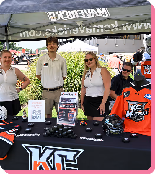 Four people standing behind a table with KC Mavericks hockey promotional items under a tent at an outdoor event.