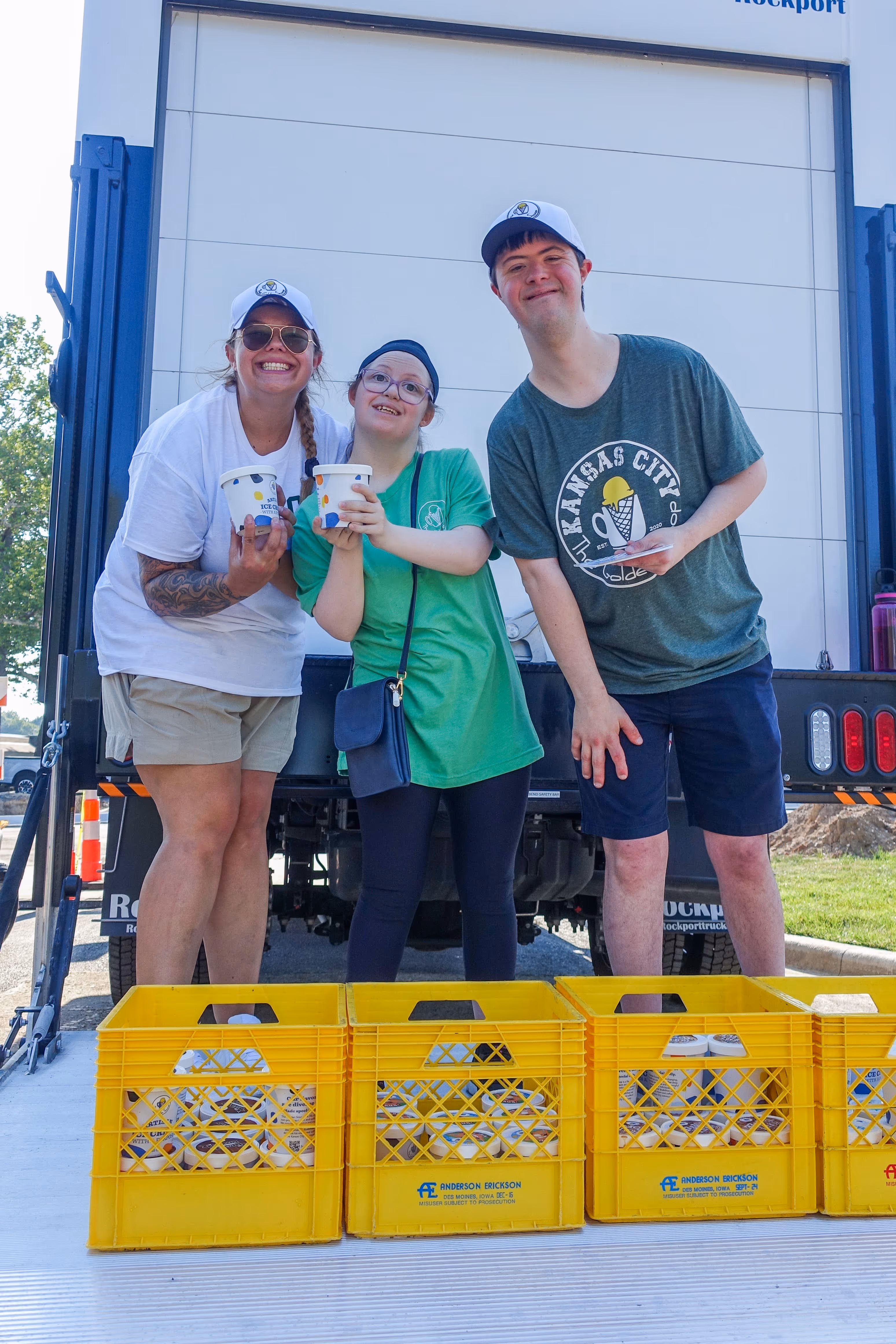 Group of people, including children and adults, gathered outdoors near a stage with some hugging and others standing nearby in casual summer clothing.