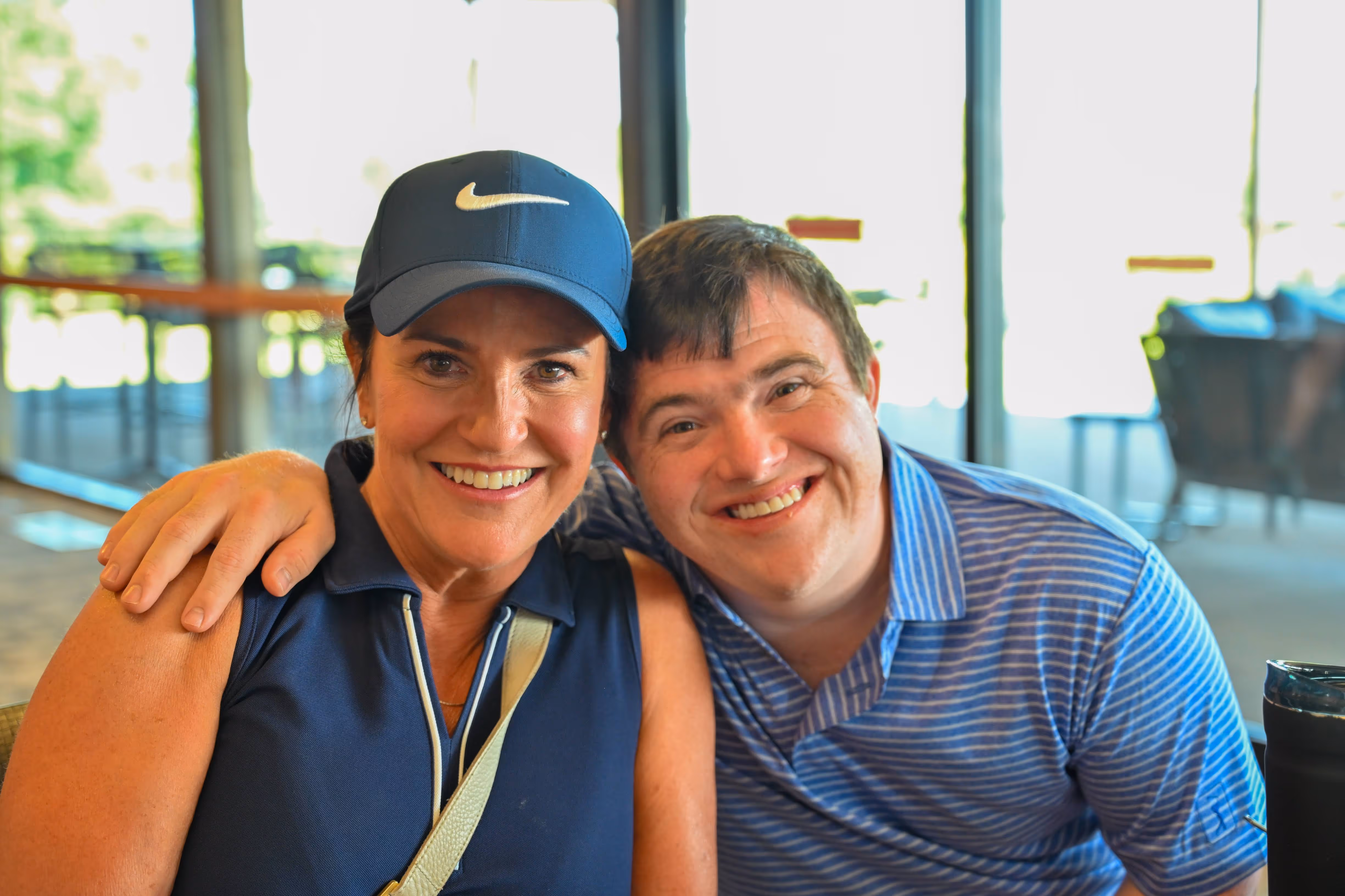 Group of people, including children and adults, gathered outdoors near a stage with some hugging and others standing nearby in casual summer clothing.