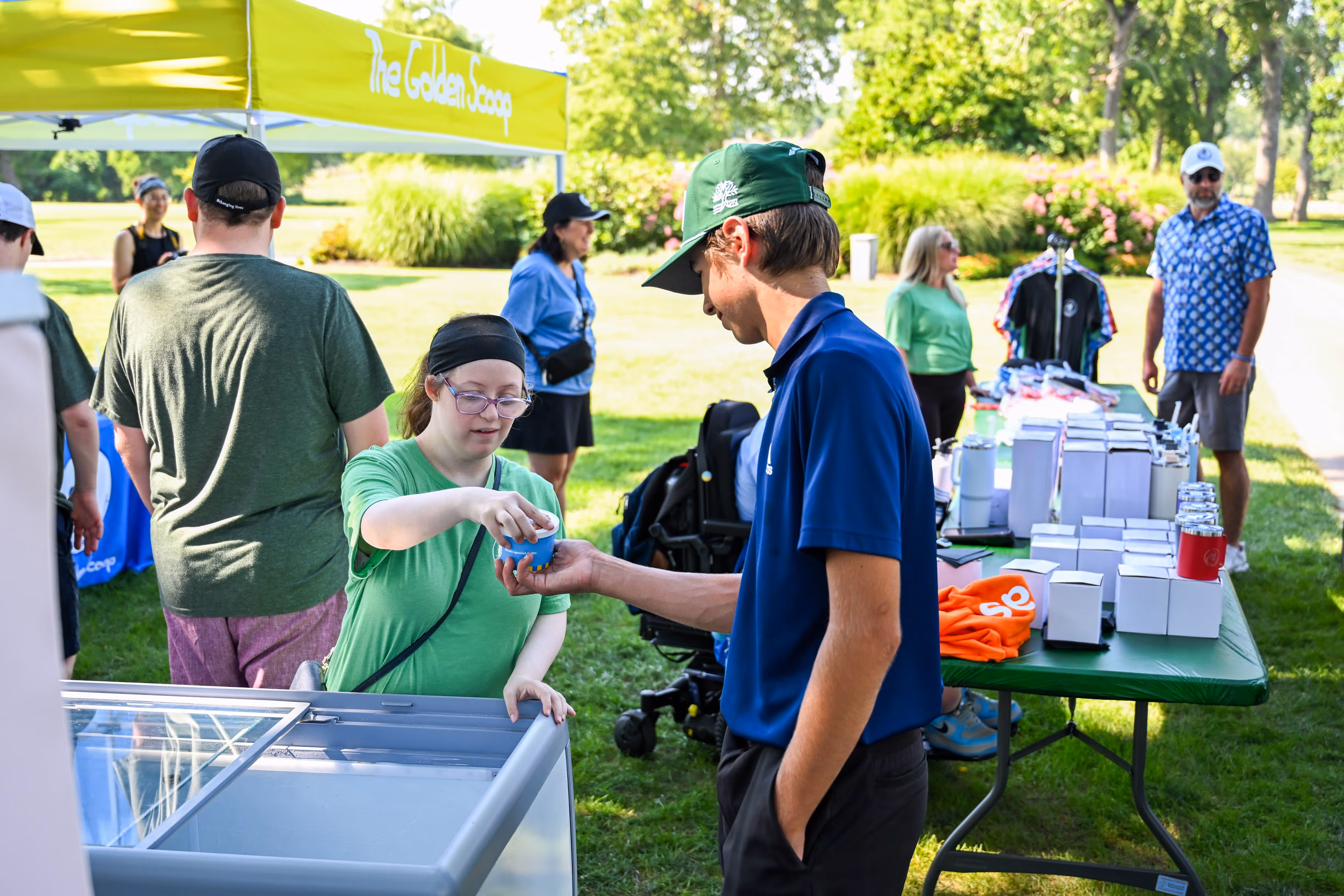 Group of people, including children and adults, gathered outdoors near a stage with some hugging and others standing nearby in casual summer clothing.