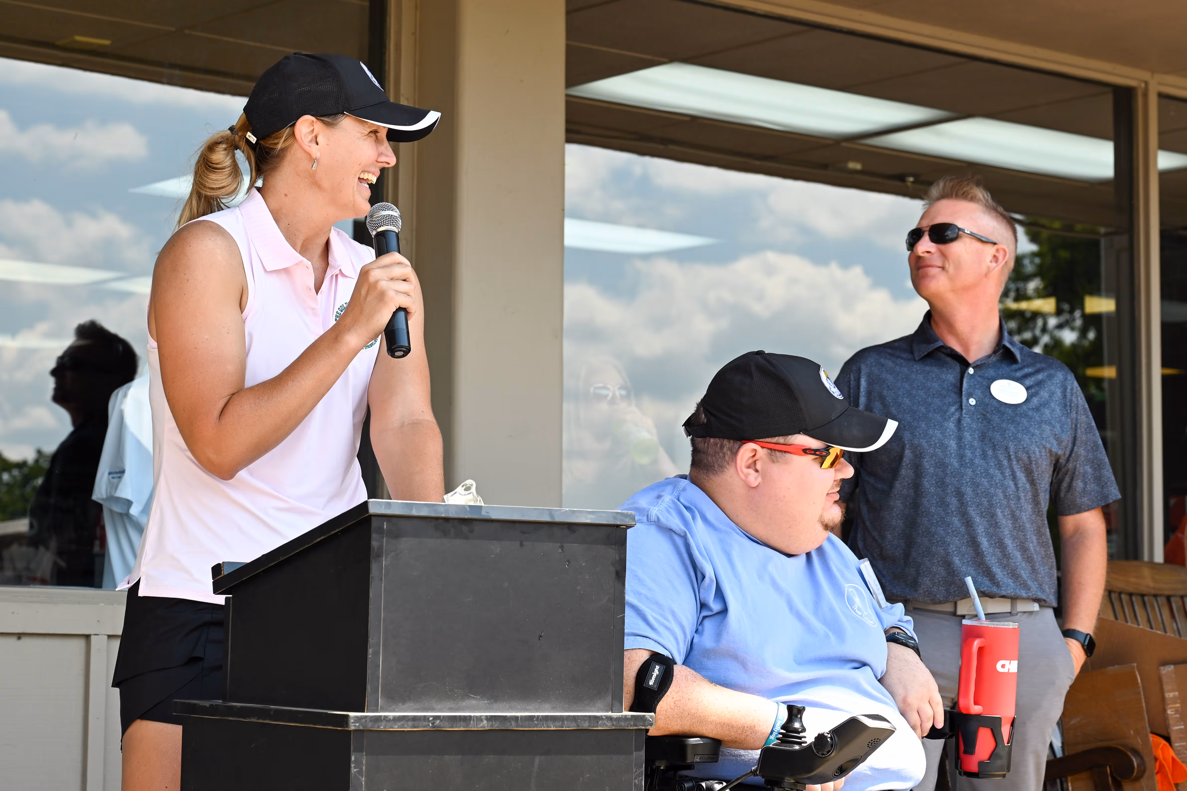 Group of people, including children and adults, gathered outdoors near a stage with some hugging and others standing nearby in casual summer clothing.