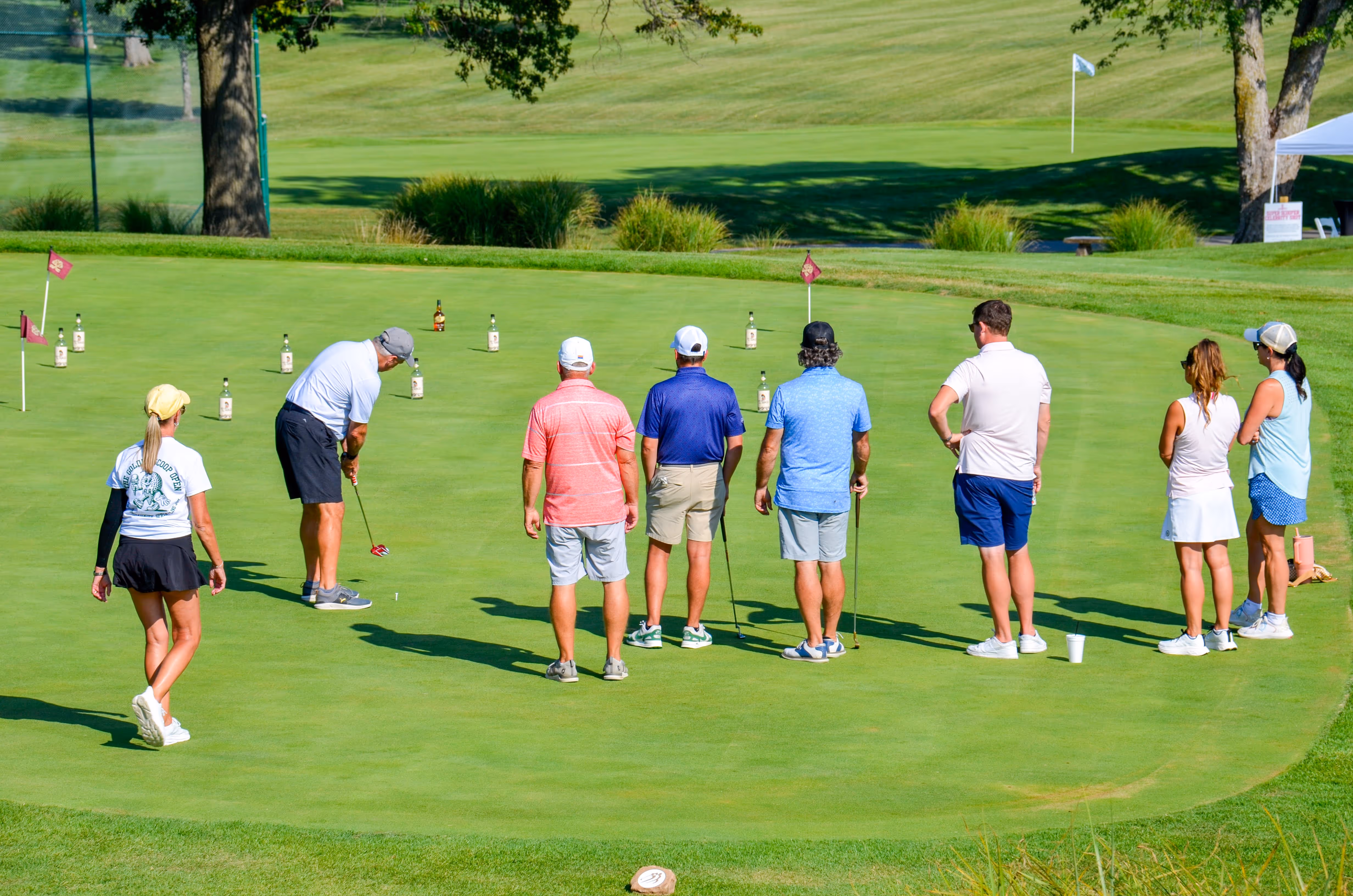 Group of people, including children and adults, gathered outdoors near a stage with some hugging and others standing nearby in casual summer clothing.