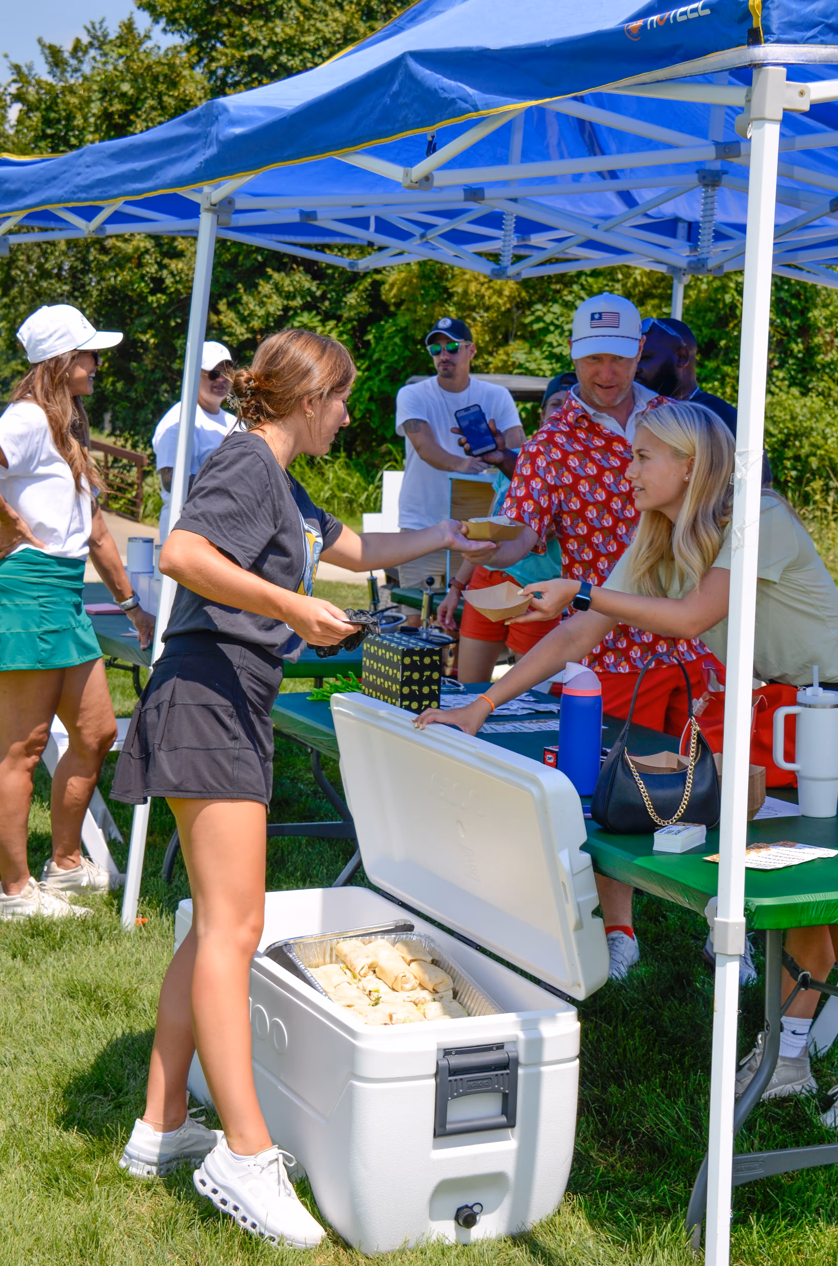 Group of people, including children and adults, gathered outdoors near a stage with some hugging and others standing nearby in casual summer clothing.